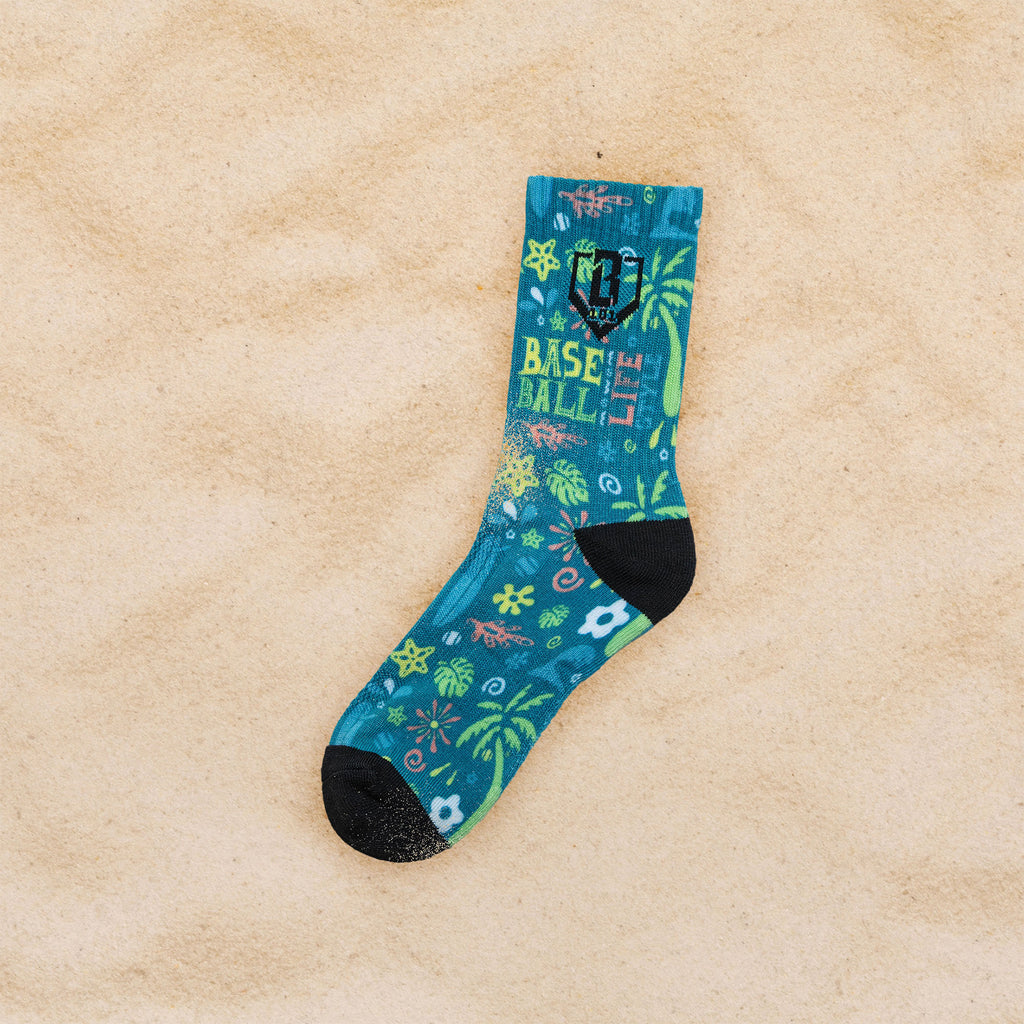 A single crew sock with a tropical beach print and 'BASEBALL LIFESTYLE' text, laid flat on a sandy beige background.