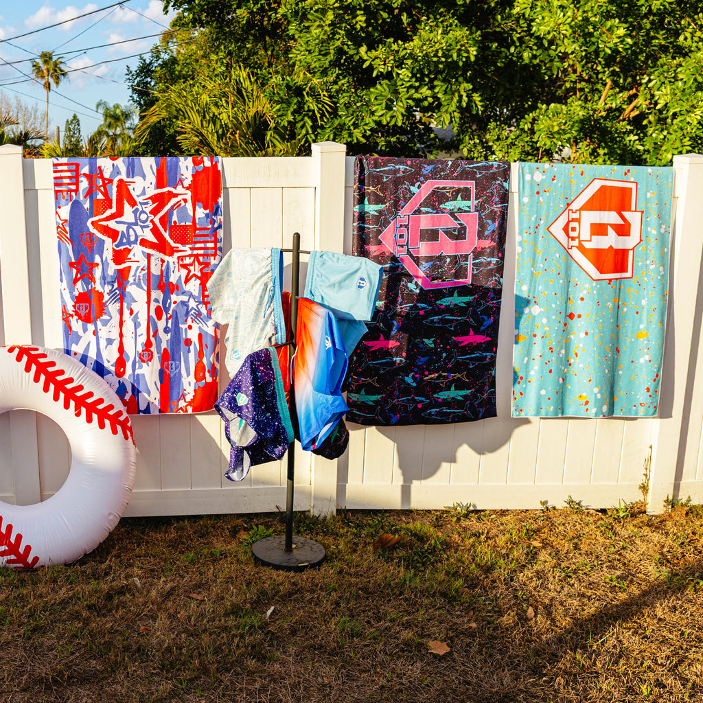 Three BL101 beach towels hanging on a white fence in graffiti, neon shark, and teal splatter prints, with BL101 swim trunks on a rack and an inflatable baseball float nearby.