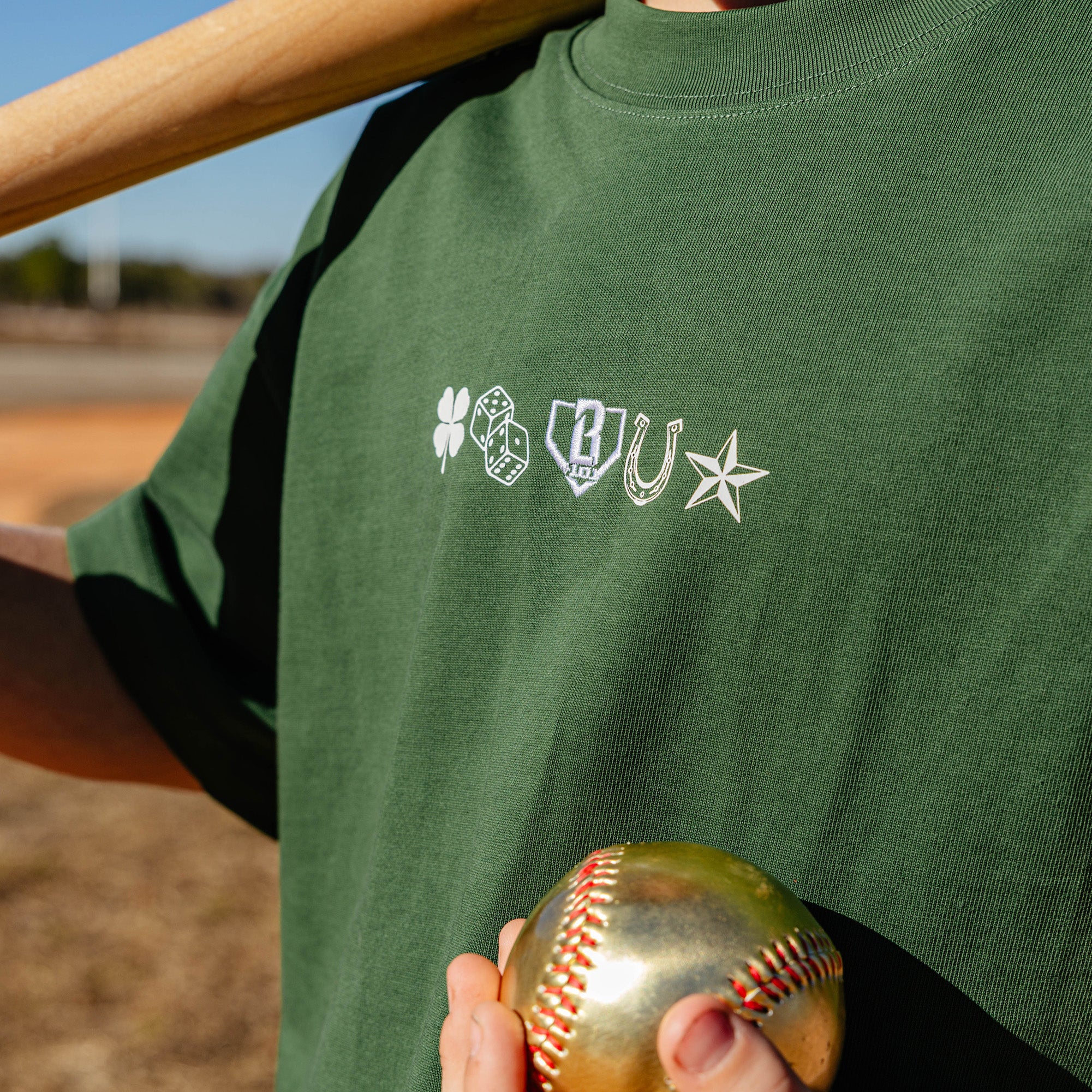 Person wearing a green t-shirt with logos holding a gold baseball on a field.