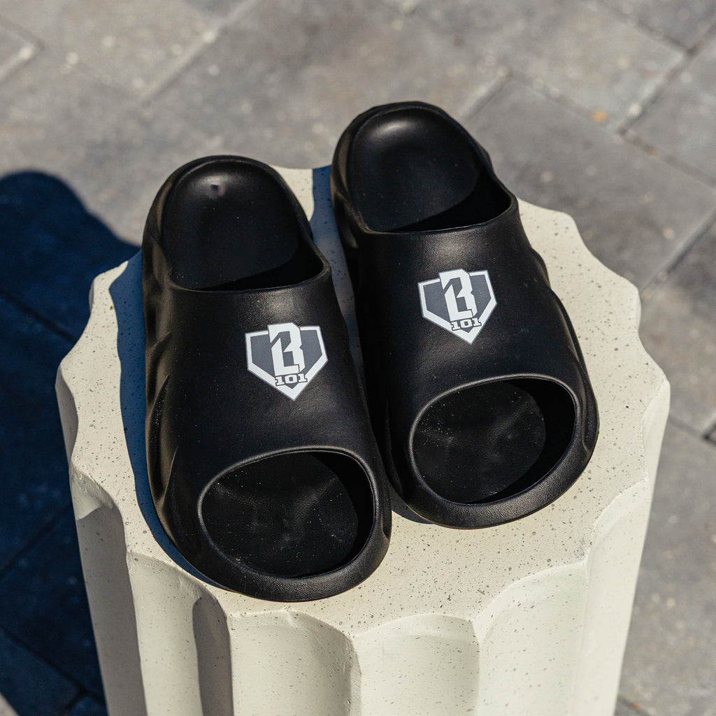 Pair of BL101 black slides displayed on a white concrete pedestal, shot from the front.