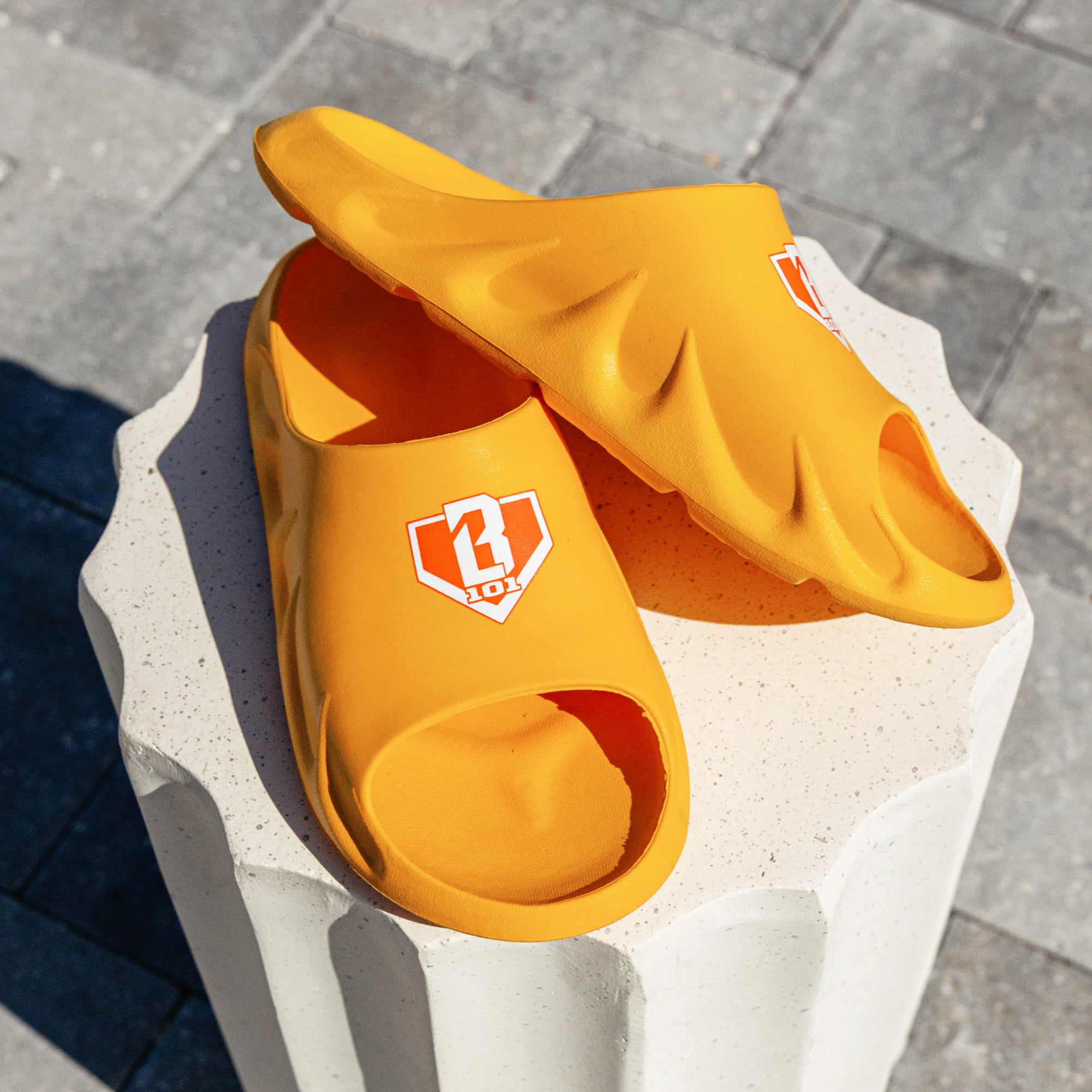 Pair of BL101 yellow slides stacked on a white concrete pedestal showing the sculpted sole and orange BL101 home plate logo.