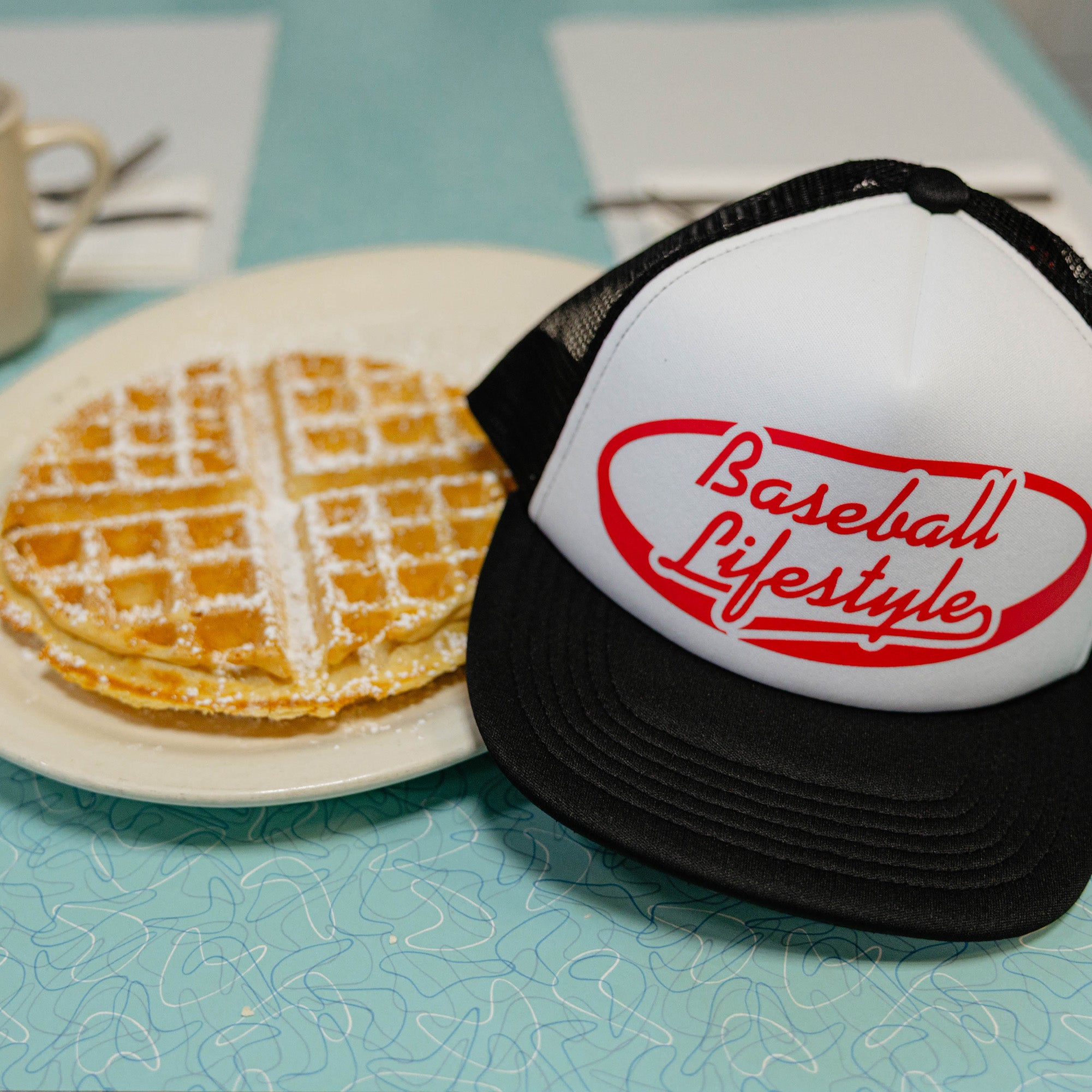 Trucker hat with 'Baseball Lifestyle' text next to waffles on a table