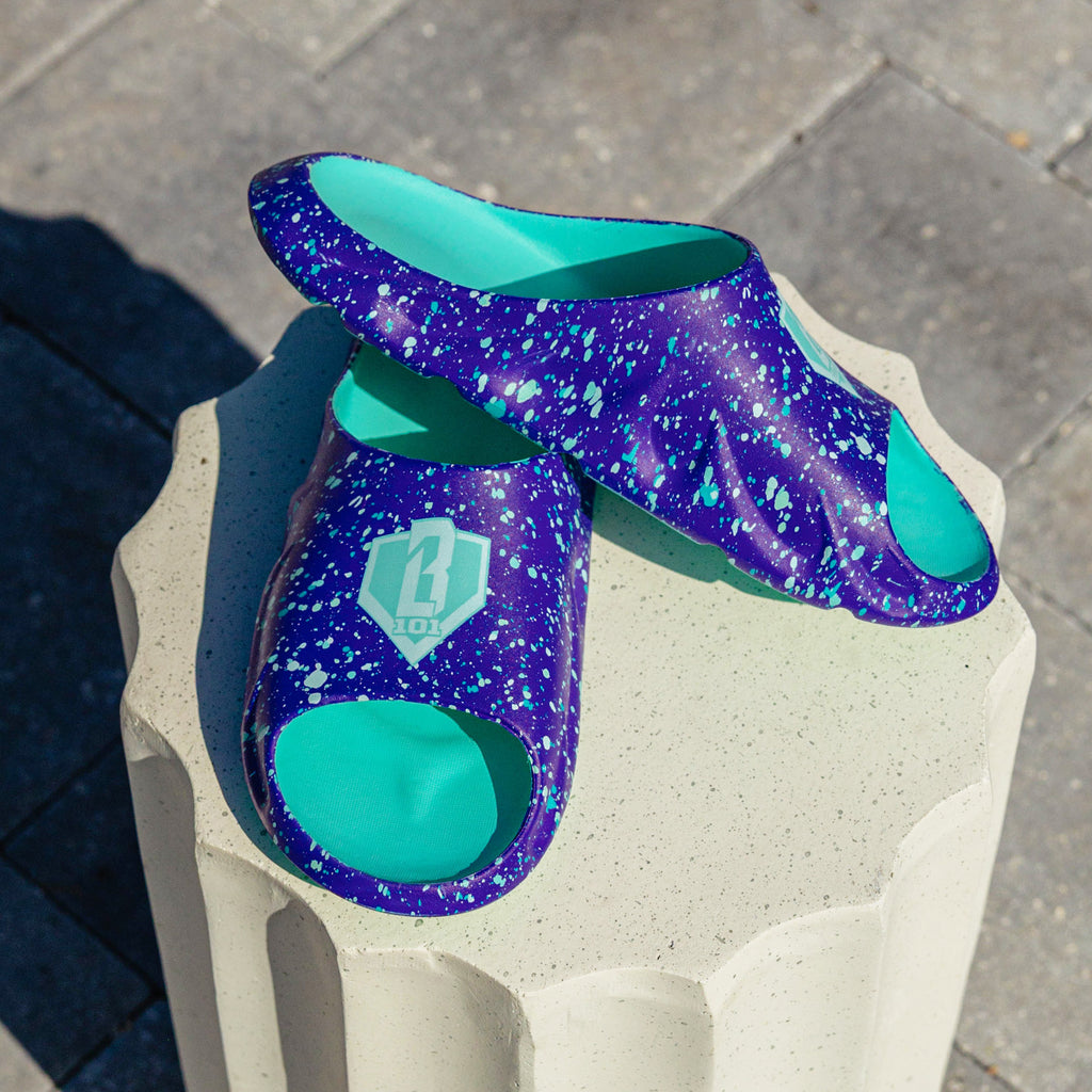 Pair of BL101 purple splatter slides stacked on a white concrete pedestal showing the sculpted sole and teal BL101 logo.
