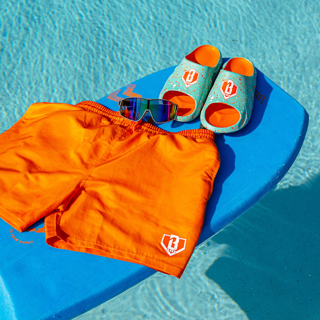 BL101 orange swim trunks and teal splatter sandals styled with shield sunglasses on a blue pool float poolside.