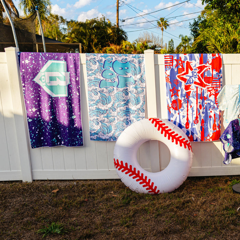 Three BL101 beach towels hanging on a white fence in purple splatter, wave, and graffiti prints, with an inflatable baseball pool float leaning against the fence.