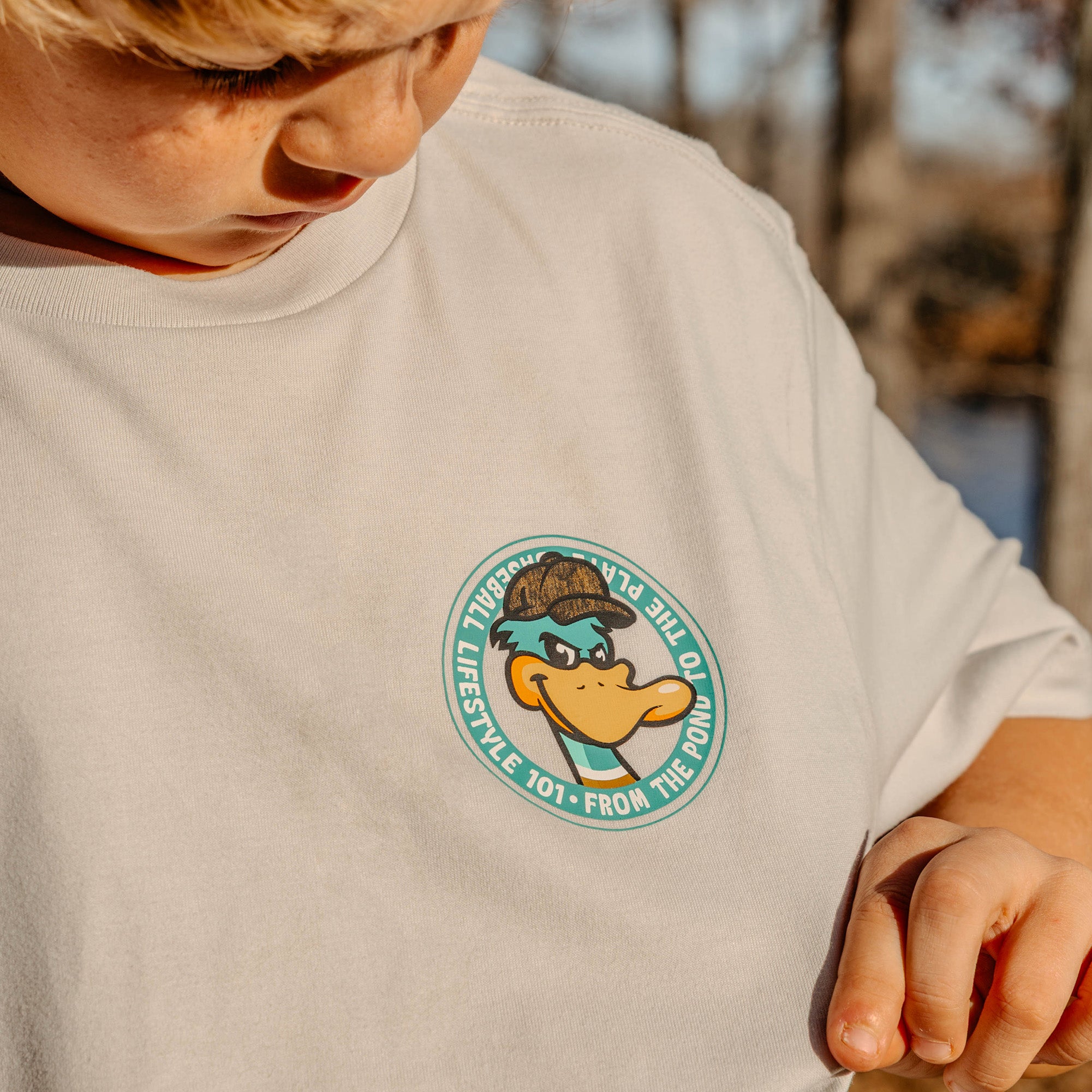 Child wearing a light gray t-shirt with a duck character logo on a blurred natural background