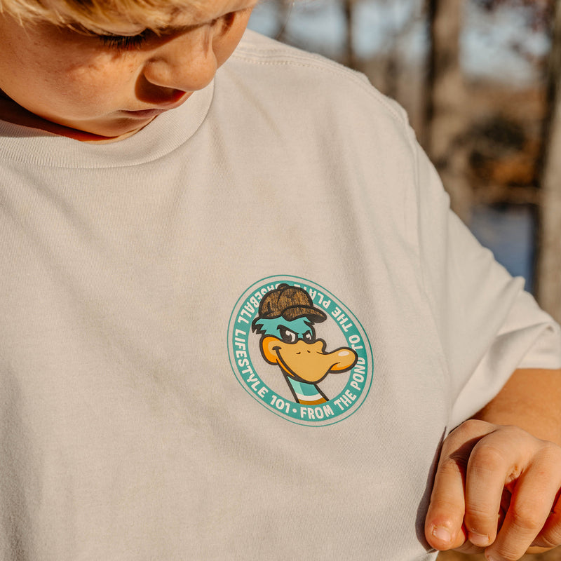 Child wearing a light gray t-shirt with a duck character logo on a blurred natural background
