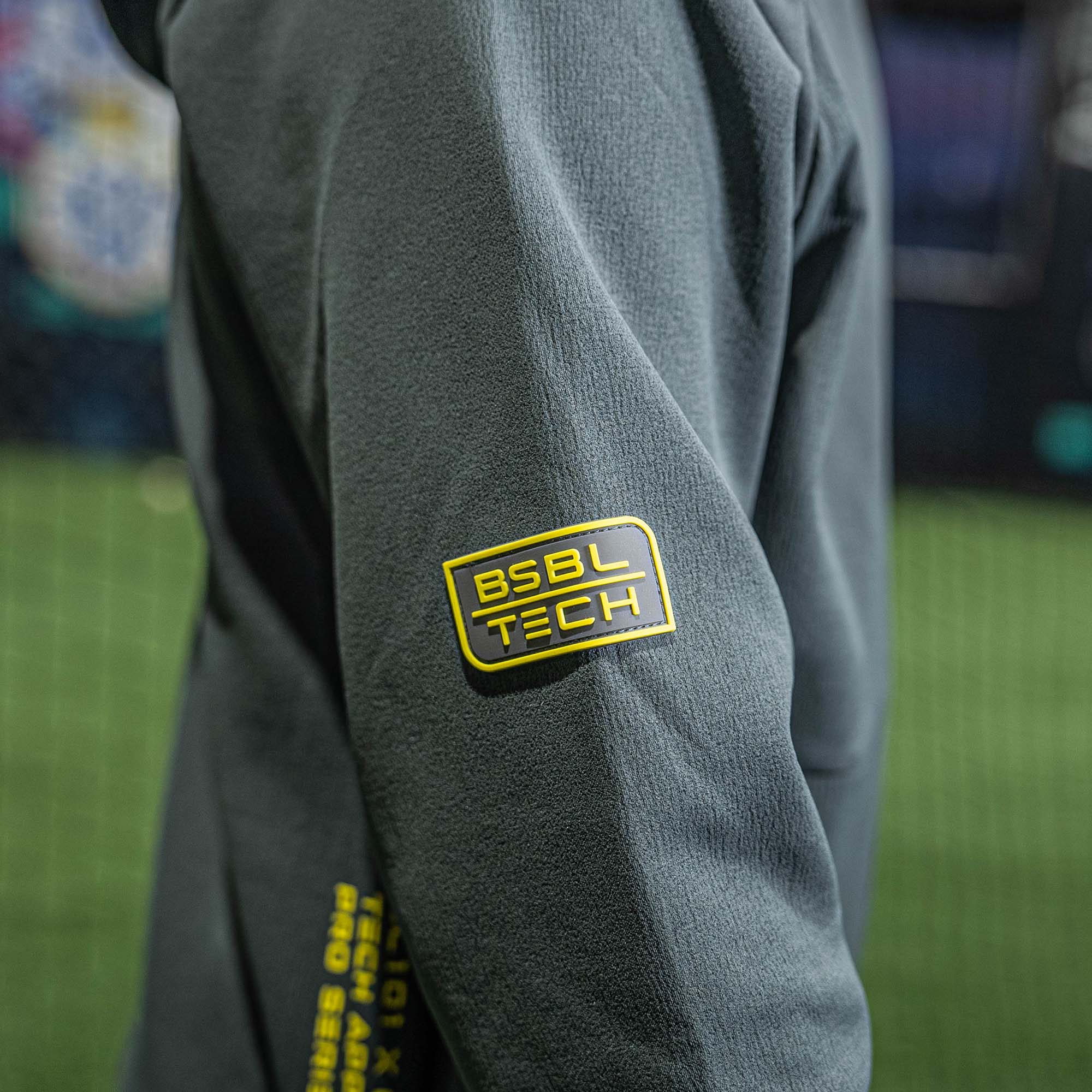 Gray fleece jacket with yellow 'BSBL Tech' logo on sleeve against a blurred sports field background