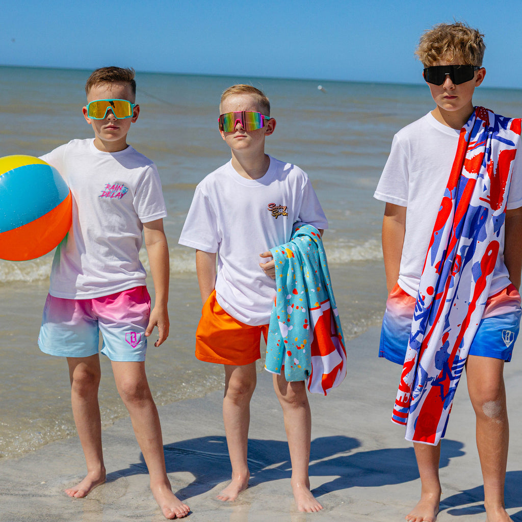 Three boys standing at the shoreline in BL101 graphic tees and swim trunks, wearing sunglasses and holding a beach ball and BL101 towels.
