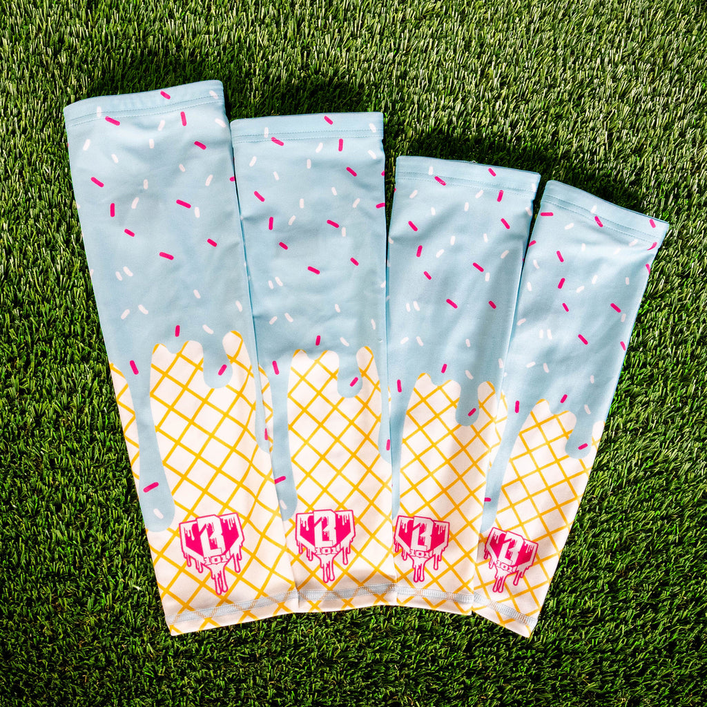 Four cotton candy ice cream arm sleeves fanned out on green turf showing the light blue sprinkle and waffle cone print with pink BL101 logos