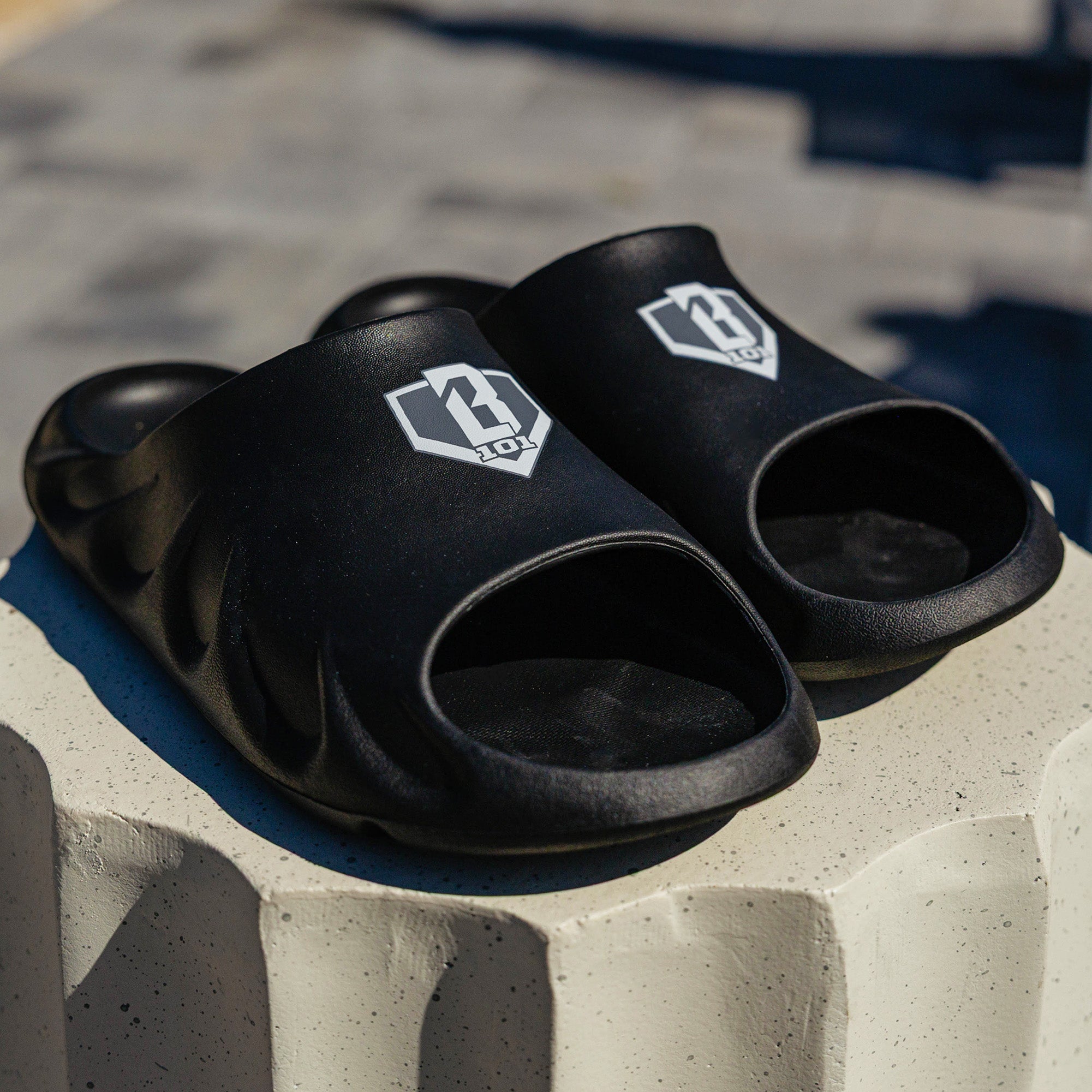 Angled shot of BL101 black slides on a white concrete pedestal with the BL101 logo visible on the strap