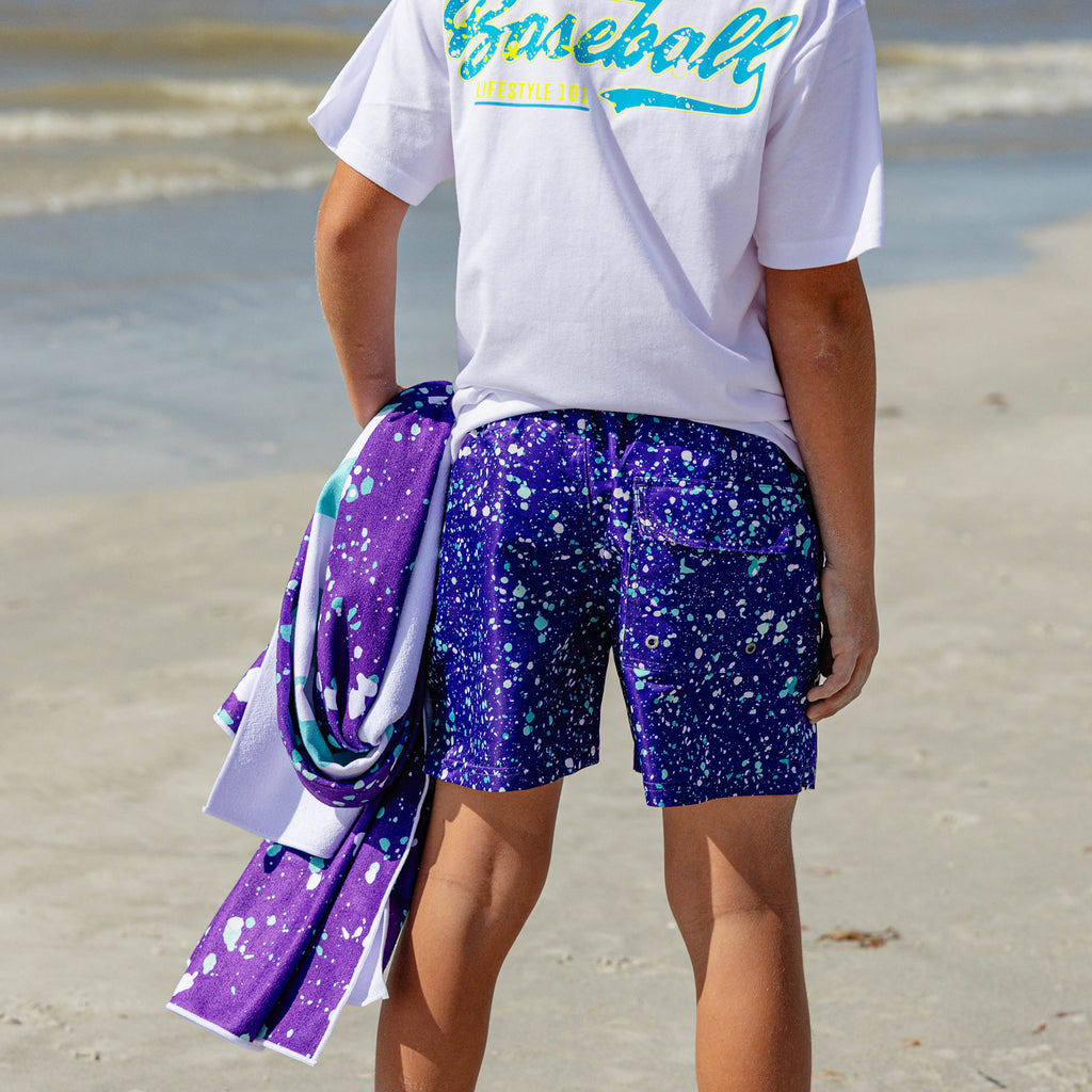 Back view of a person on the beach wearing BL101 purple splatter swim trunks and a Baseball Lifestyle 101 tee, holding a matching purple splatter towel.