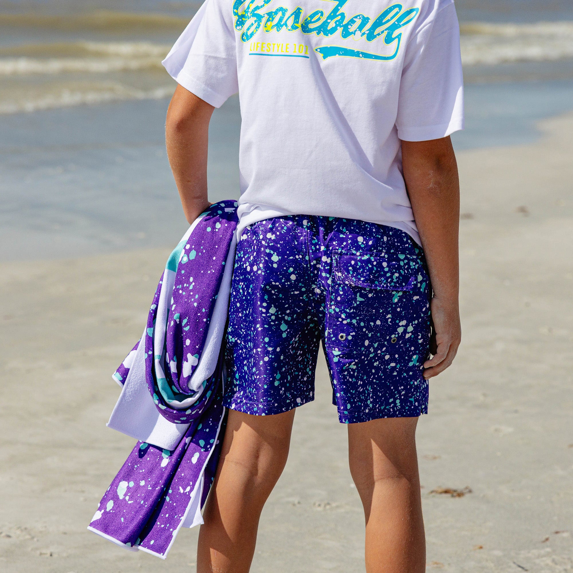 Back view of a person on the beach wearing BL101 purple splatter swim trunks and a Baseball Lifestyle 101 tee, holding a matching purple splatter towel.