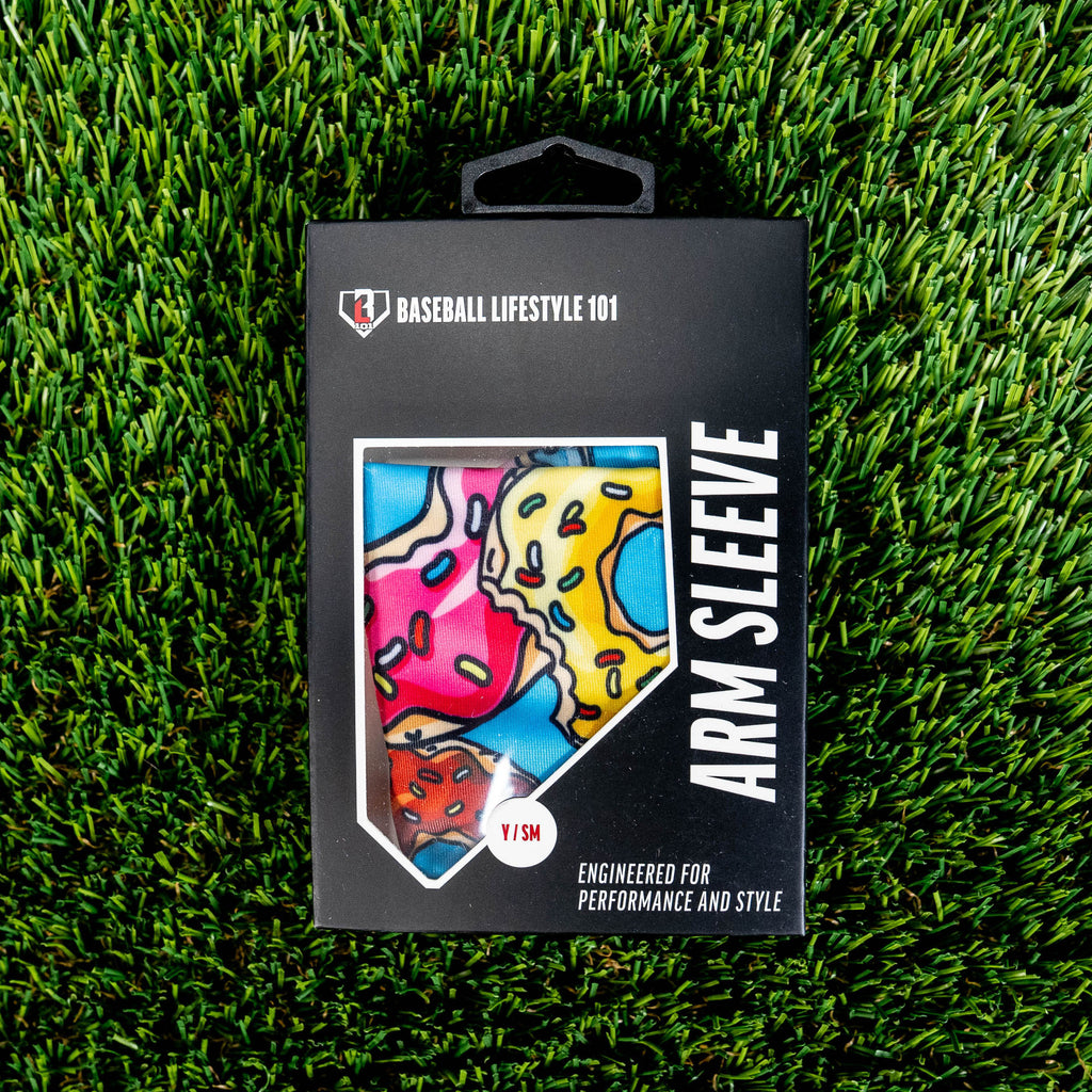Front of the BL101 arm sleeve retail box on green turf showing the donut print through a home plate window and Y/SM size label