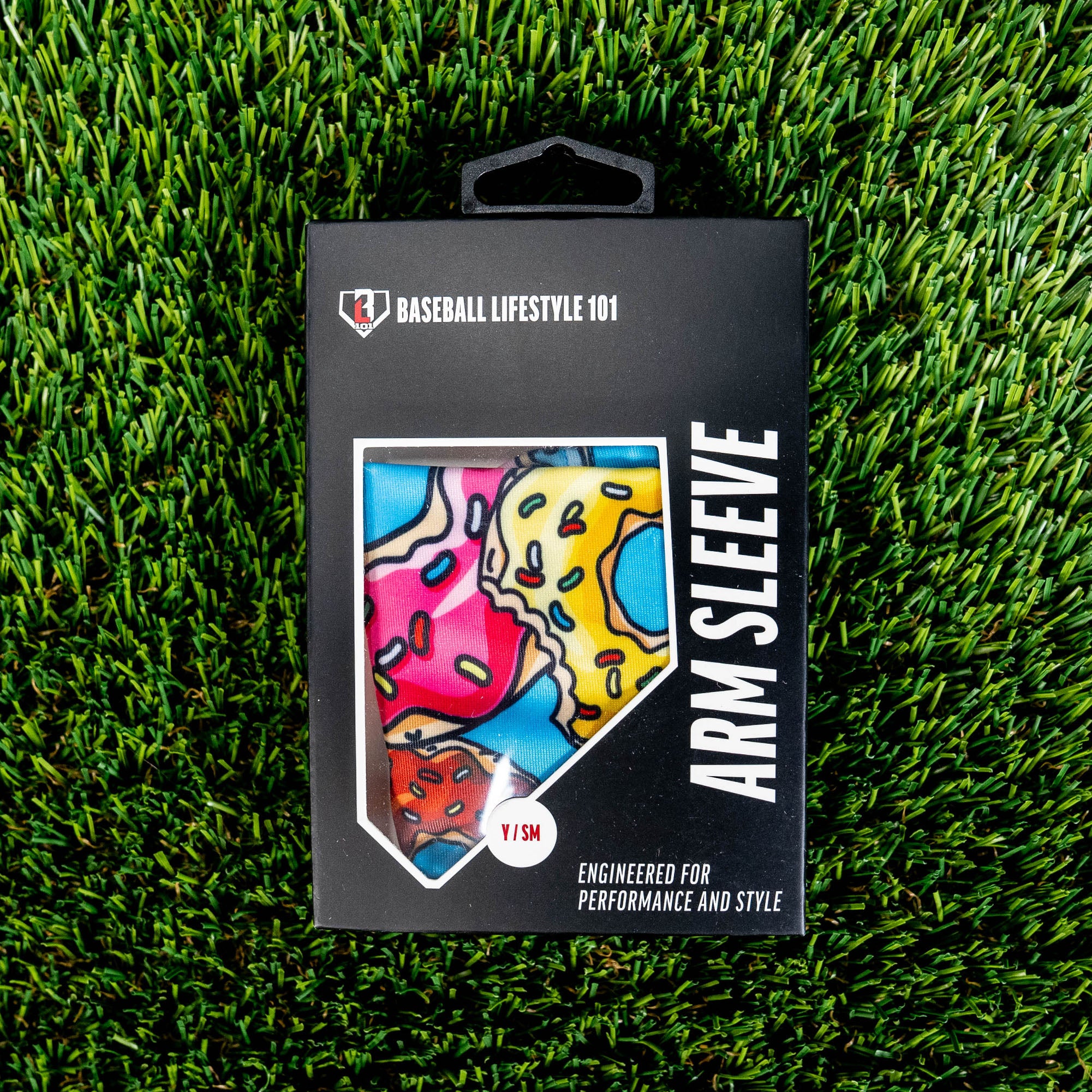 Front of the BL101 arm sleeve retail box on green turf showing the donut print through a home plate window and Y/SM size label