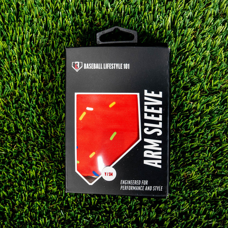 Front of the BL101 arm sleeve retail box on green turf showing the red sprinkle print through a home plate window and Y/SM size label