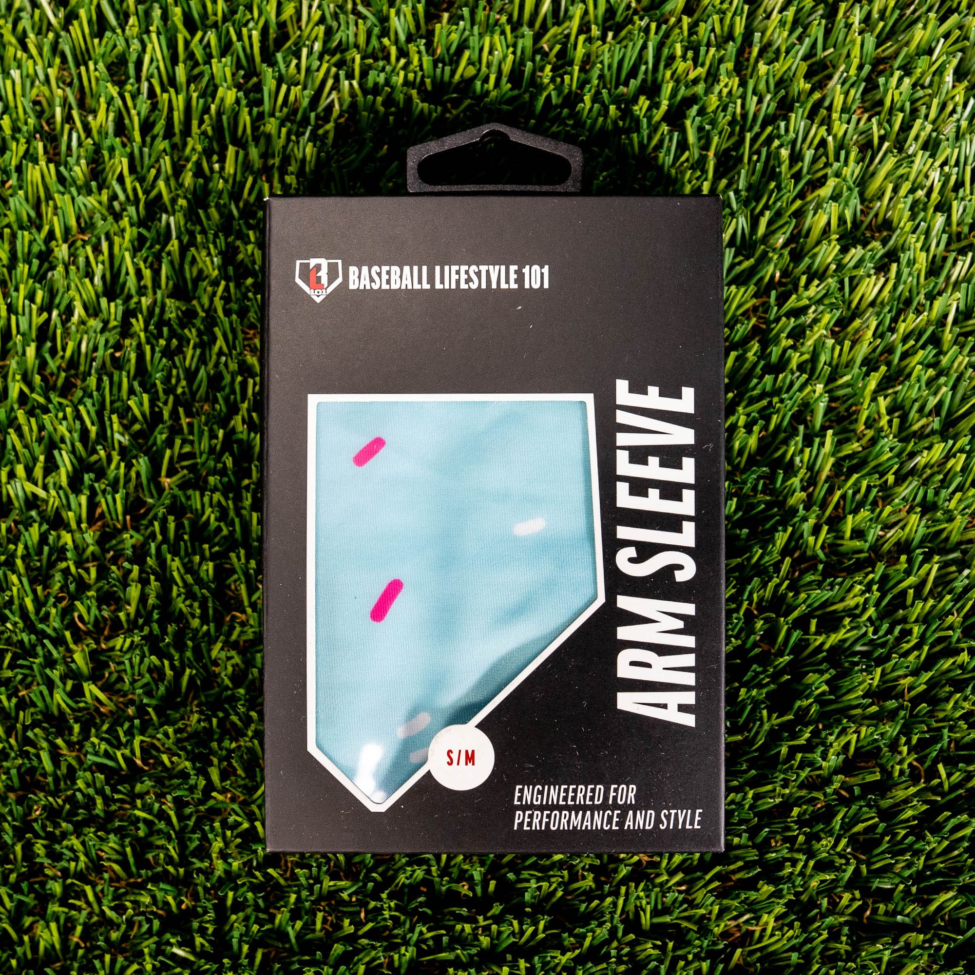 Front of the BL101 arm sleeve retail box on green turf showing the light blue sprinkle print through a home plate window and S/M size label