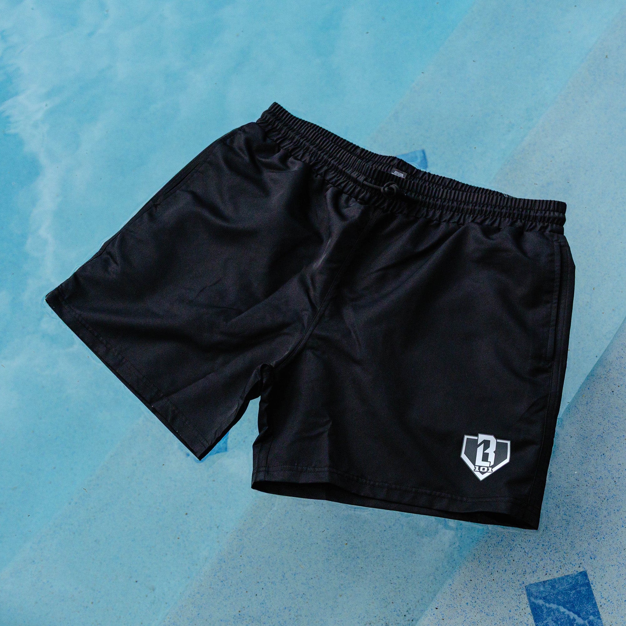 Black BL101 swim trunks with elastic drawstring waistband and gray BL101 home plate logo, laid flat on a light blue pool