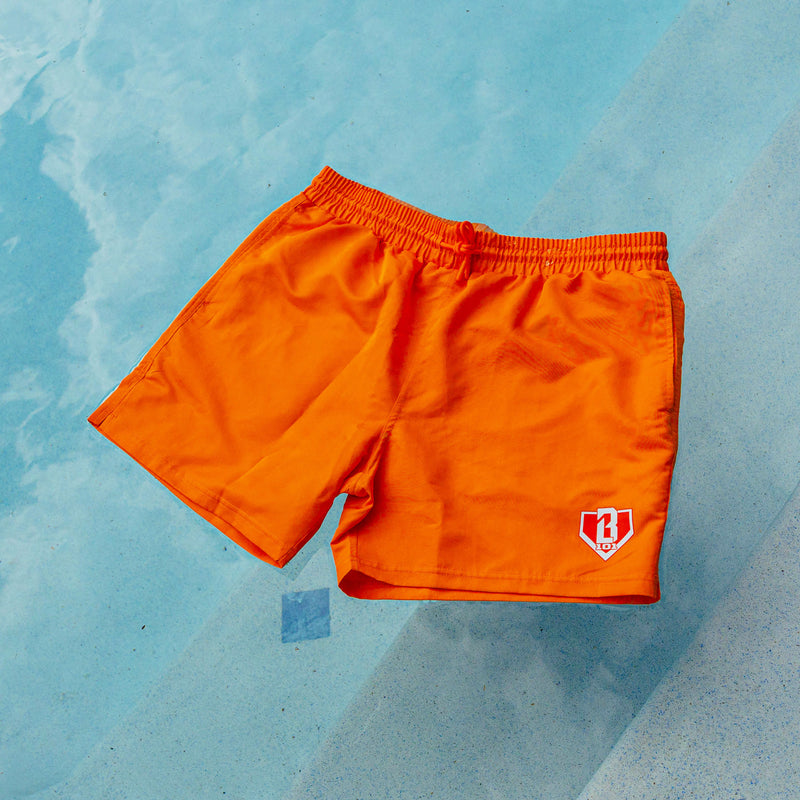 Orange BL101 swim trunks with elastic drawstring waistband and white BL101 home plate logo, laid flat on a light blue pool deck.
