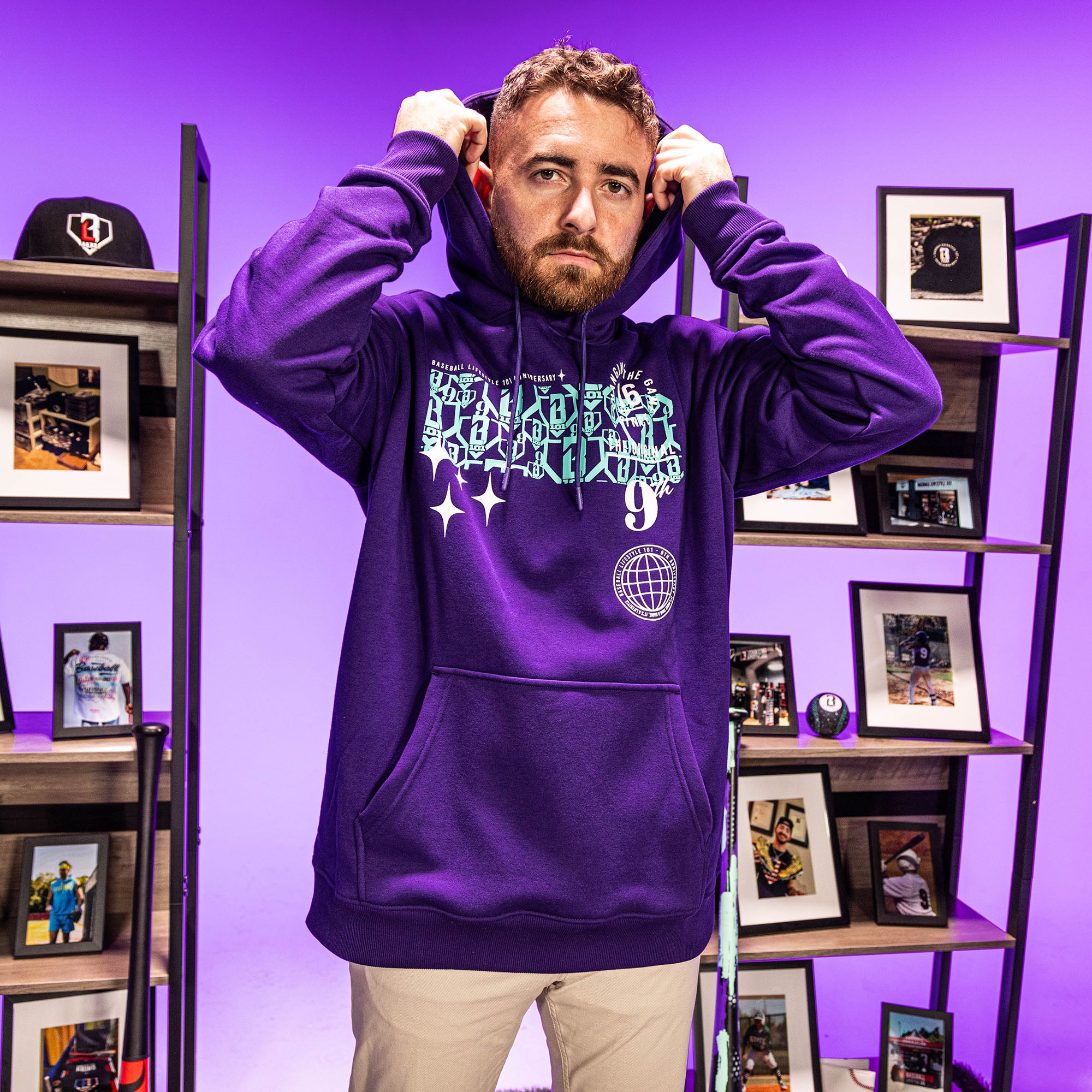 Man wearing a purple hoodie with blue BL101 9th anniversary graphic designs in a room with shelves and framed pictures.