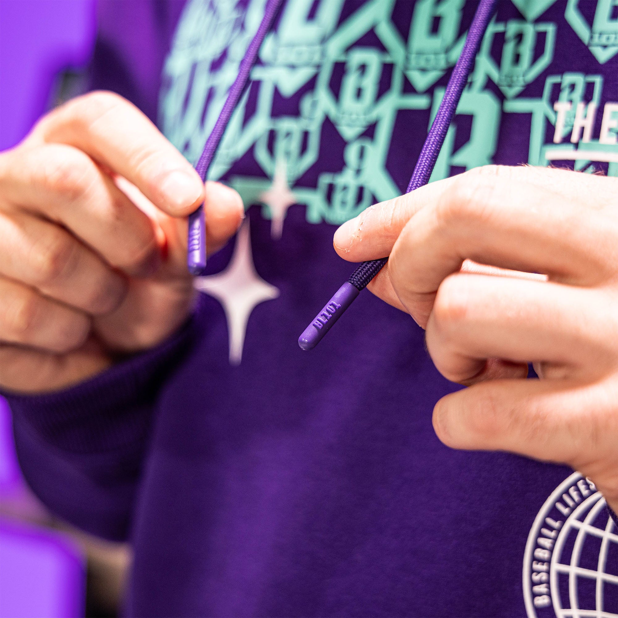 Person wearing a purple hoodie with multiple BL101 logos, holding purple strings with BL101 text on it.