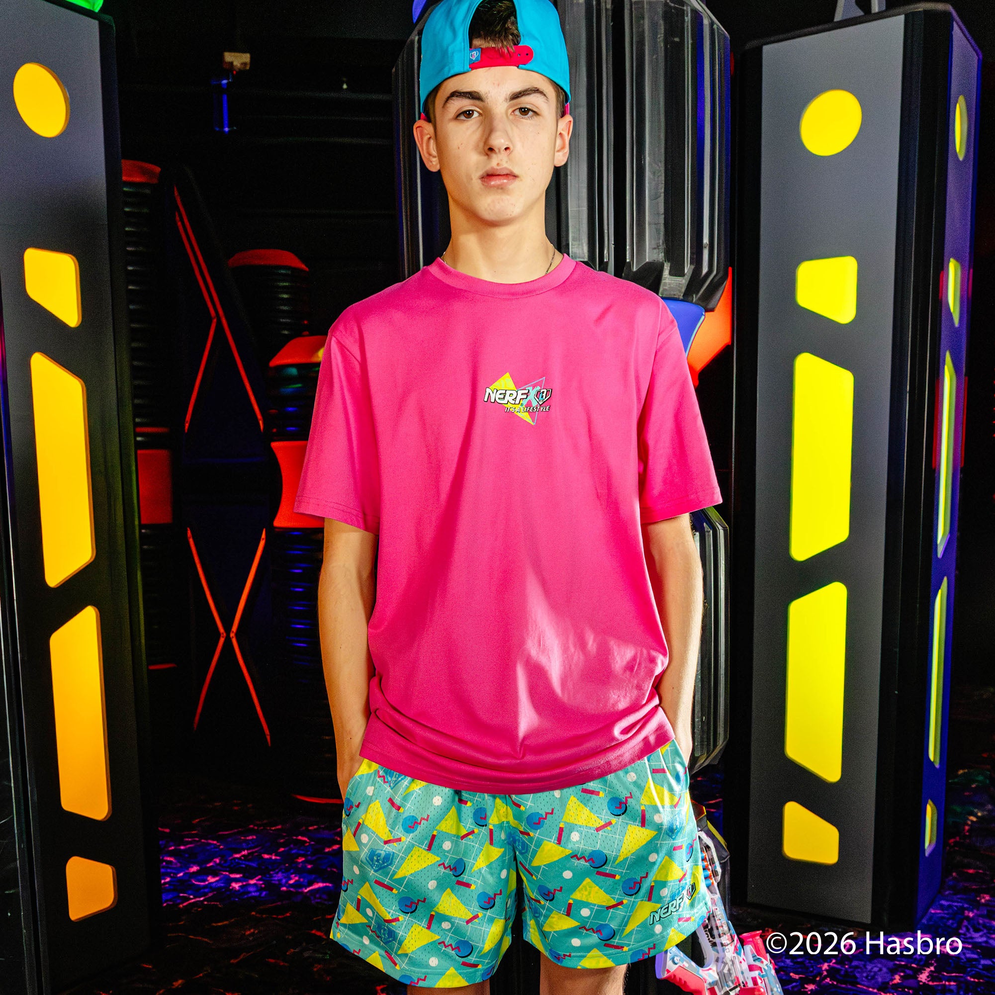 Person wearing a pink 'NERF X BL101' shirt and colorful shorts standing in front of a vibrant, futuristic background.