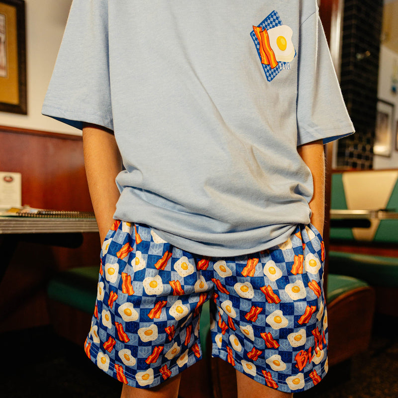 Person wearing a light blue t-shirt and patterned shorts with bacon and egg on it in a casual setting.