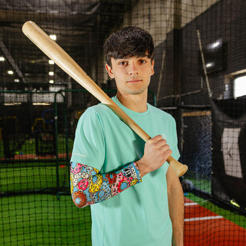 Male baseball player in mint green shirt wearing donut arm sleeve holding a wooden bat in an indoor batting cage
