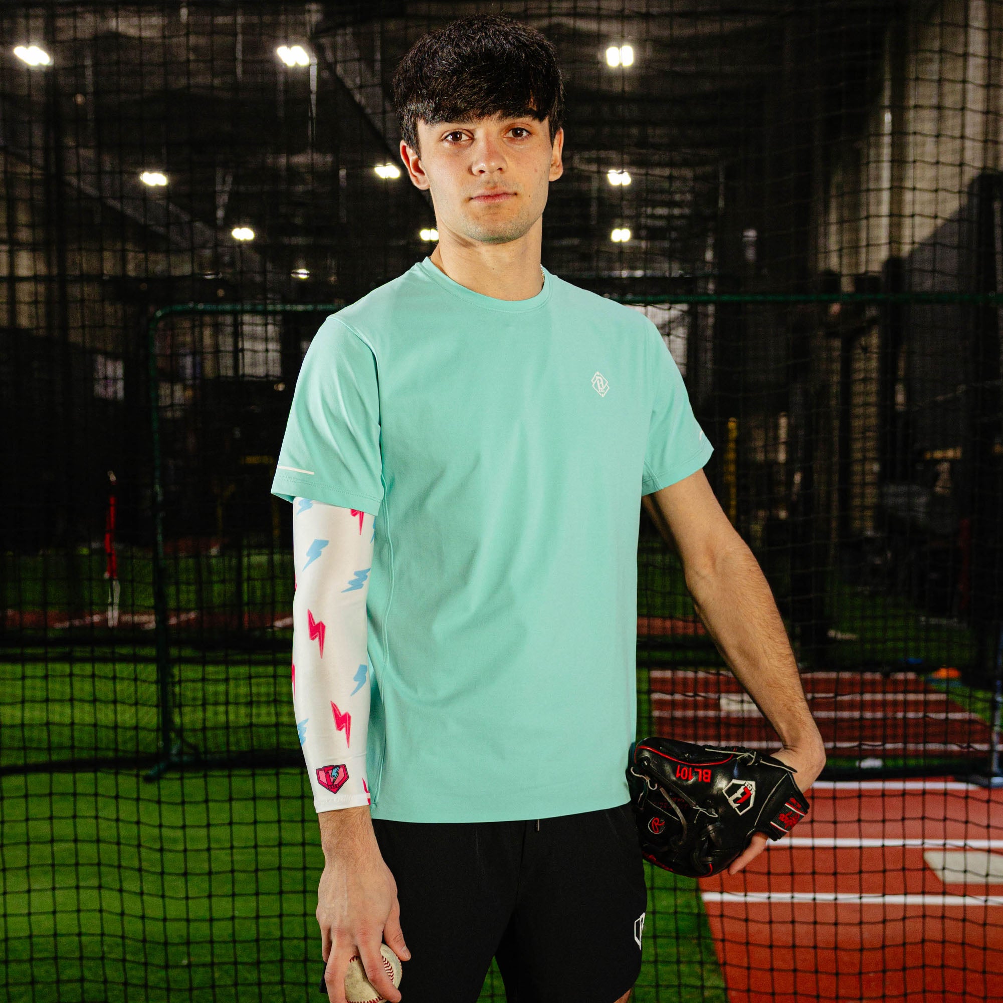 Male baseball player in a mint green shirt wearing the lightning arm sleeve on his left arm while holding a glove and baseball in an indoor batting cage