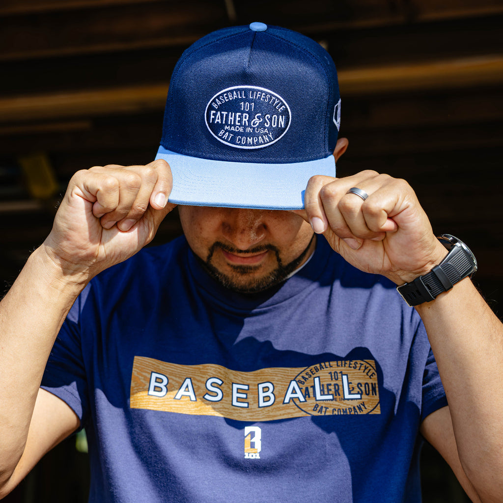 Man pulling a navy and columbia blue Father & Son Bat Company snapback down over his face while wearing a matching BL101 Father & Son baseball tee.