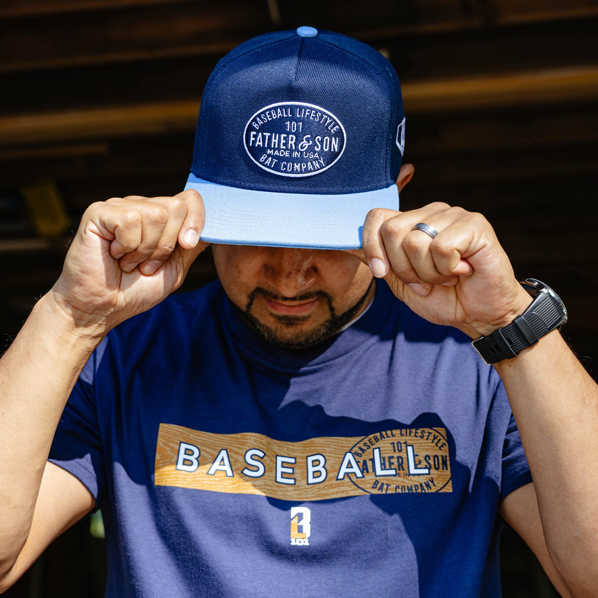 Man pulling a navy and columbia blue Father & Son Bat Company snapback down over his face while wearing a matching BL101 Father & Son baseball tee.