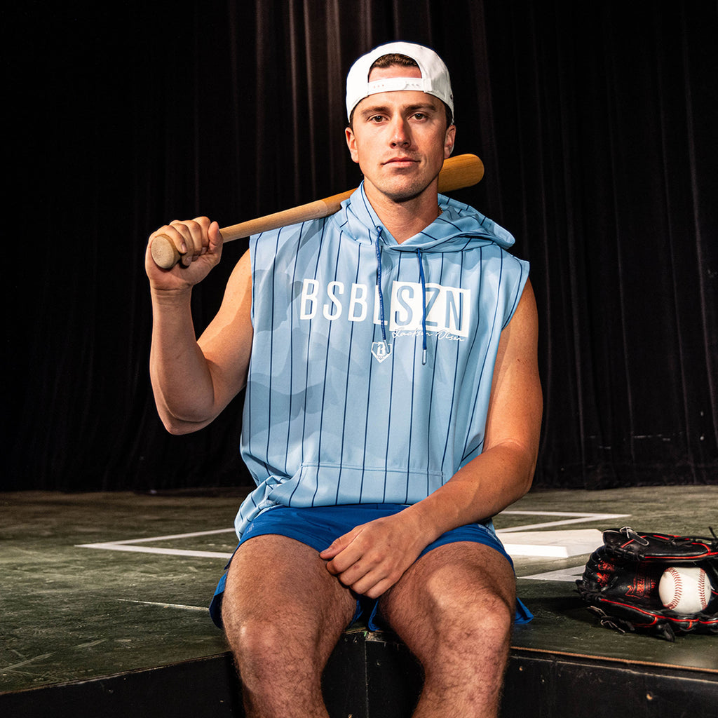Adult male model seated on a stage holding a bat over his shoulder, wearing the light blue pinstripe Jackson Olson x BL101 BSBL-SZN Short Sleeve Hoodie V3 with blue shorts