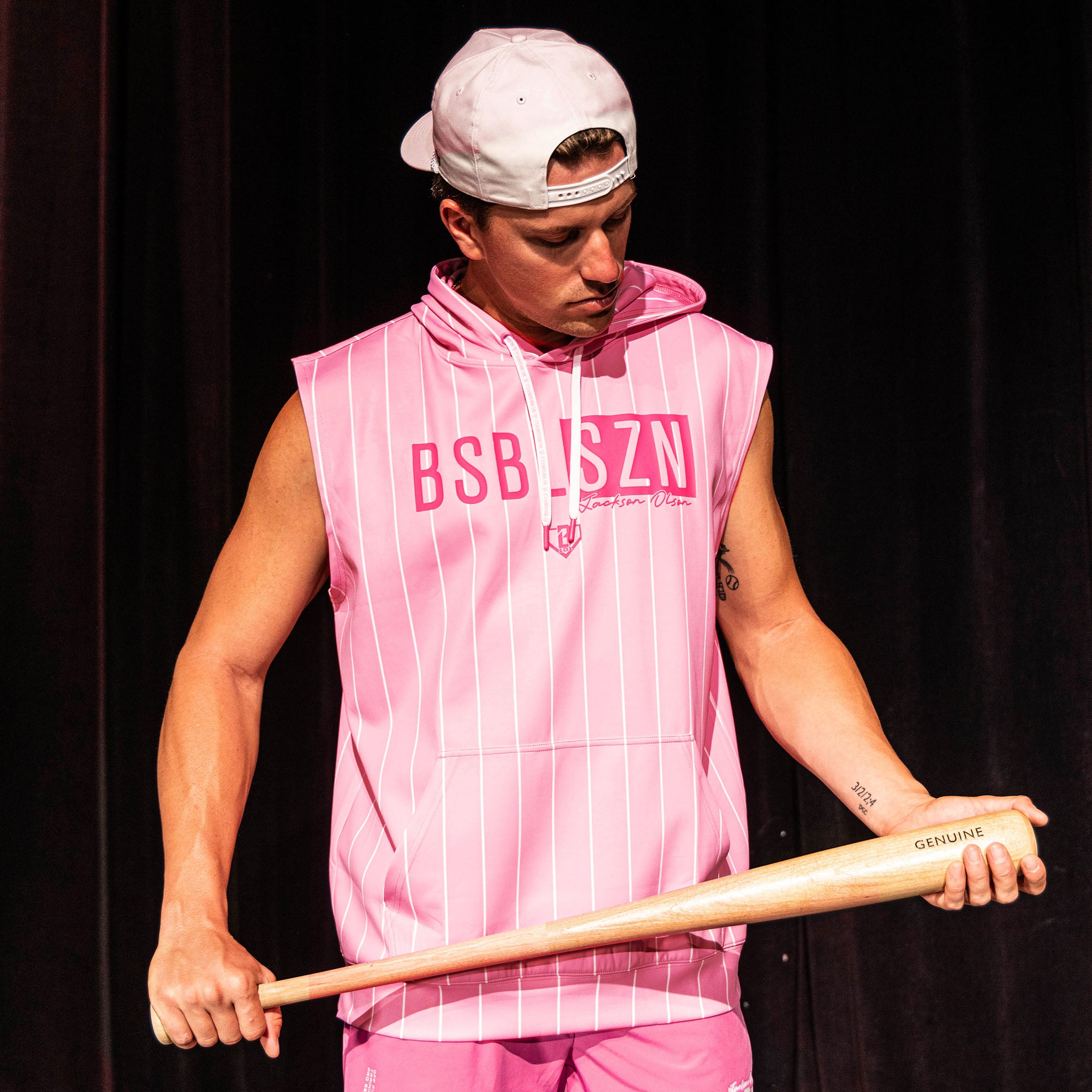 Adult male model holding a wood bat wearing the pink pinstripe Jackson Olson x BL101 BSBL-SZN Short Sleeve Hoodie V3 with matching pink shorts