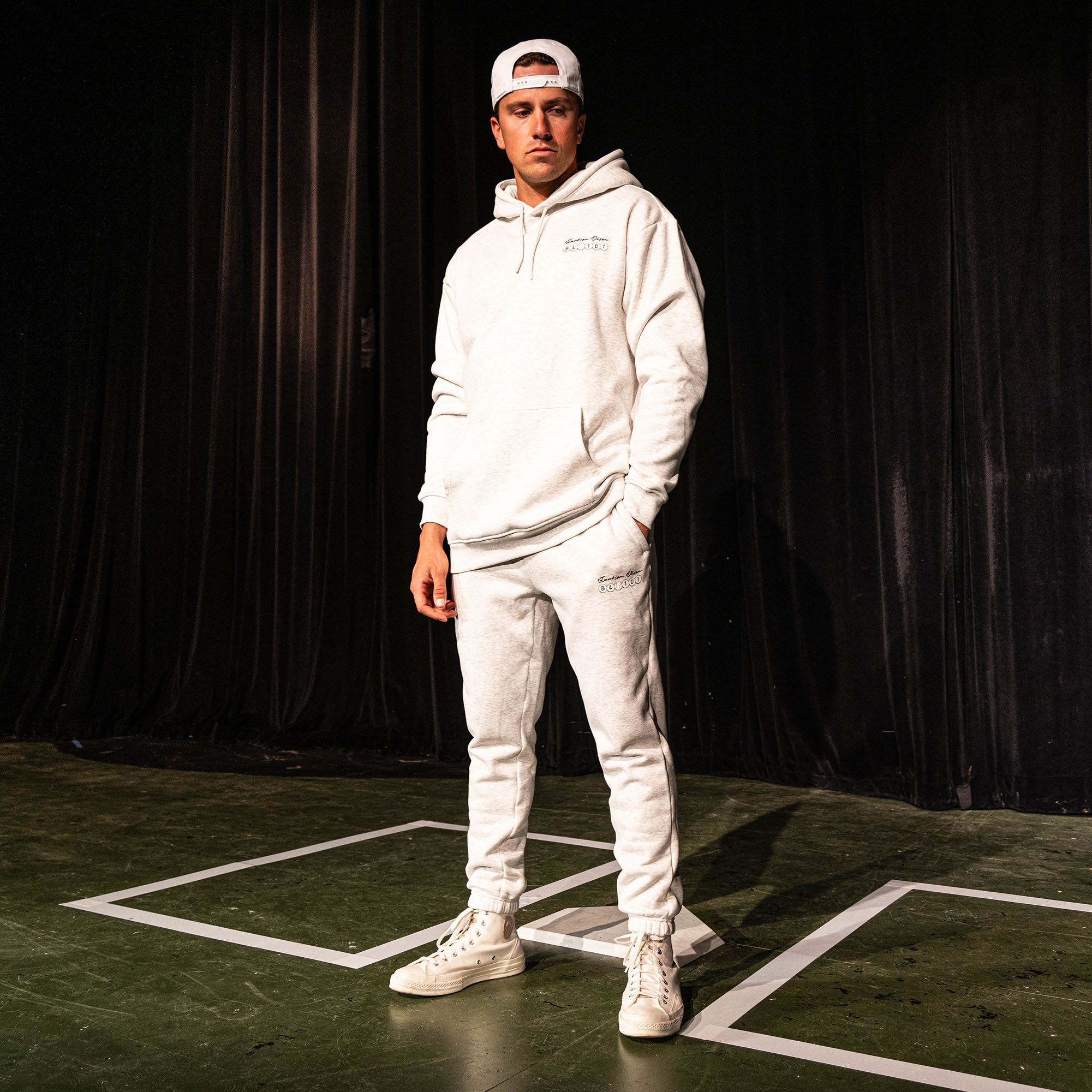 Adult male model wearing the heather gray Jackson Olson x BL101 BSBL Era sweatpants and matching hoodie, shown full-length on a stage set