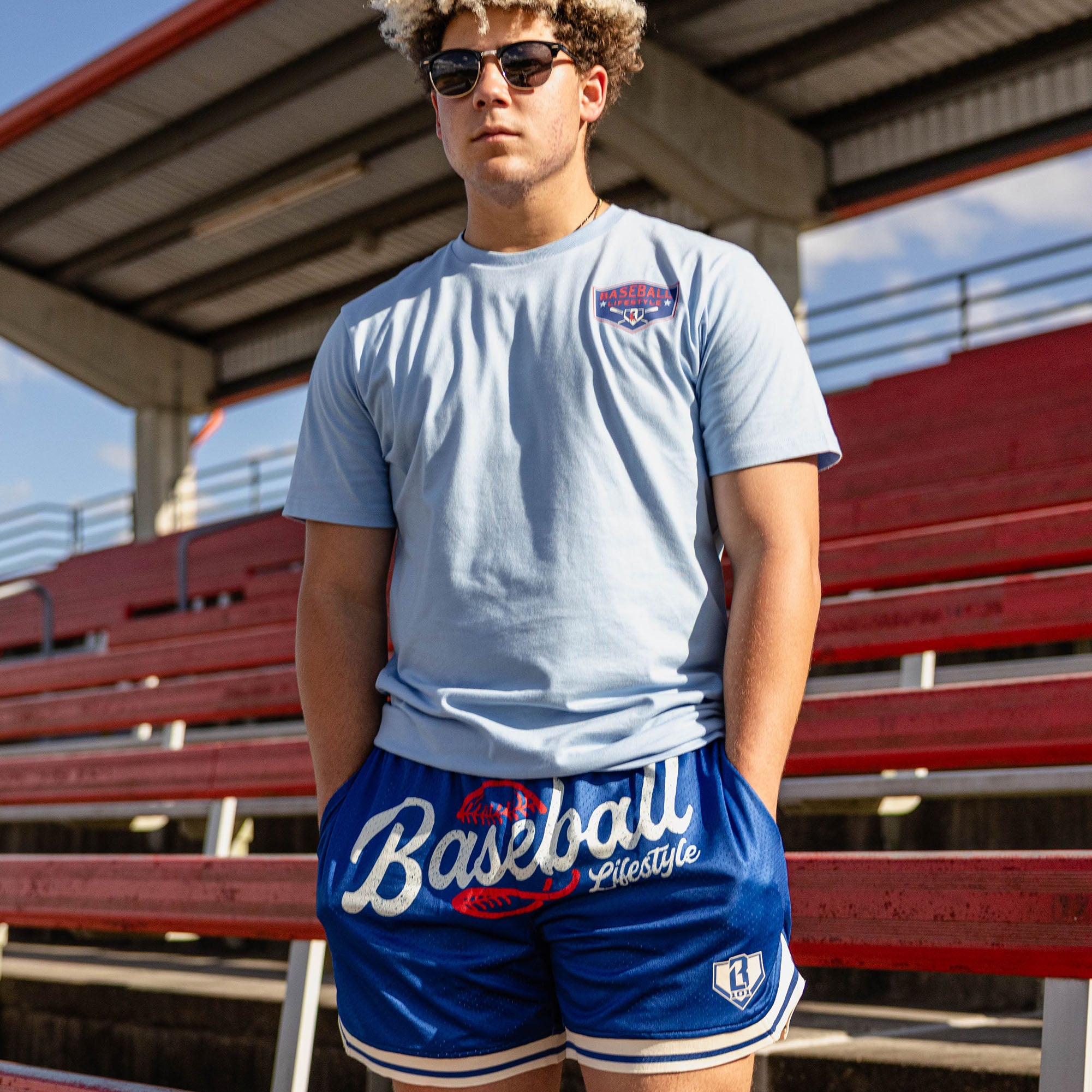 Man with curly hair in sunglasses standing at red stadium bleachers with hands in pockets wearing a light blue tee with a left chest BL101 logo graphic and royal blue Baseball Lifestyle mesh shorts