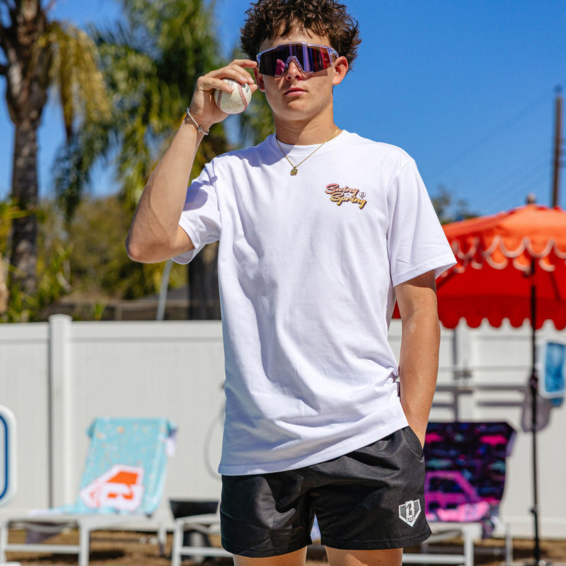  Male model standing poolside in black BL101 swim trunks and a white Swing Into Spring tee, holding a baseball while wearing pink mirrored shield sunglasses.