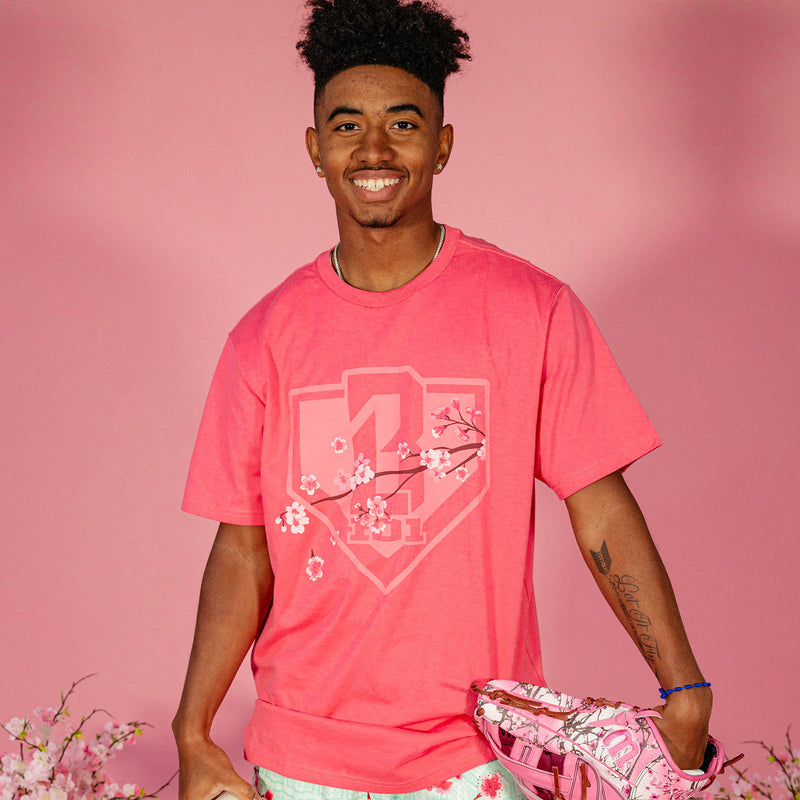 Male model wearing a pink BL101 cherry blossom shield tee and holding a pink floral baseball glove against a pink background, front view