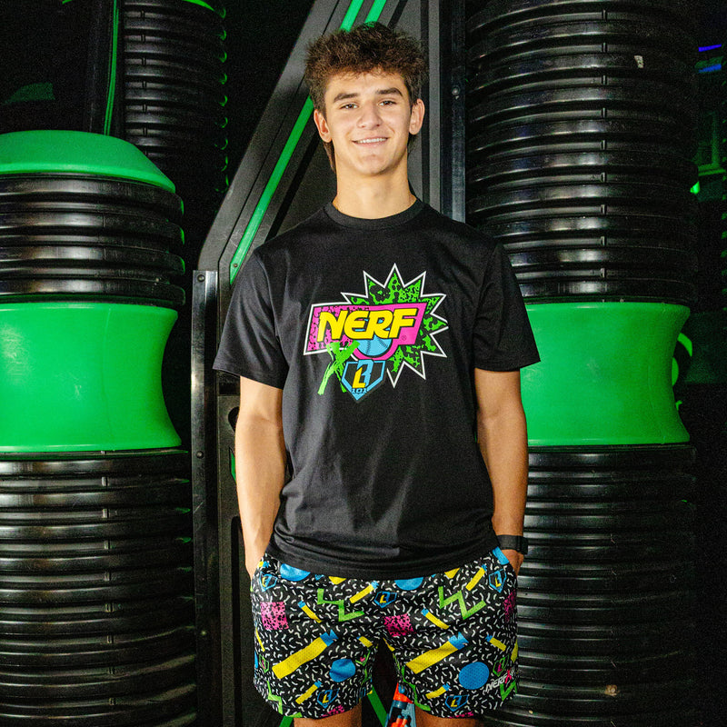 Person wearing a black t-shirt with a colorful 'NERF X BL101' logo and nerf dart patterned shorts standing in front of green and black barrels.