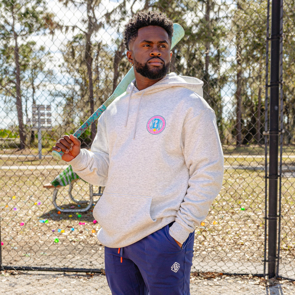 Man holding a baseball bat, wearing a light gray hoodie with a colorful BL101 logo and blue pants, standing in an outdoor setting.
