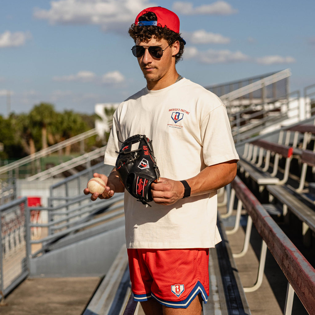 Man holding a baseball and glove at stadium bleachers wearing a red snapback aviator sunglasses a cream tee and red mesh shorts with blue and white striped hem