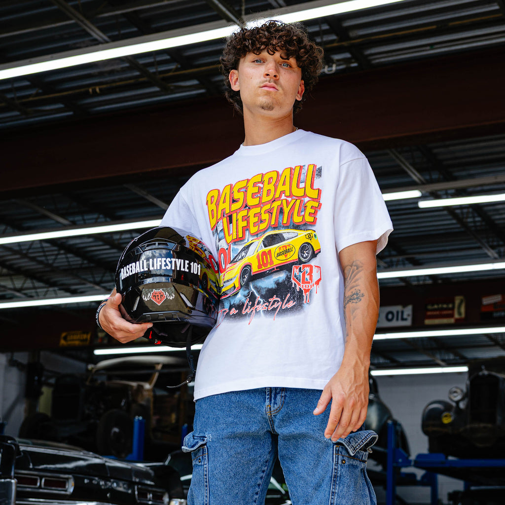 Young man holding racing helmet in vintage garage, wearing white Diamond Drift tee with yellow and red BL101 race car graphic and jeans.