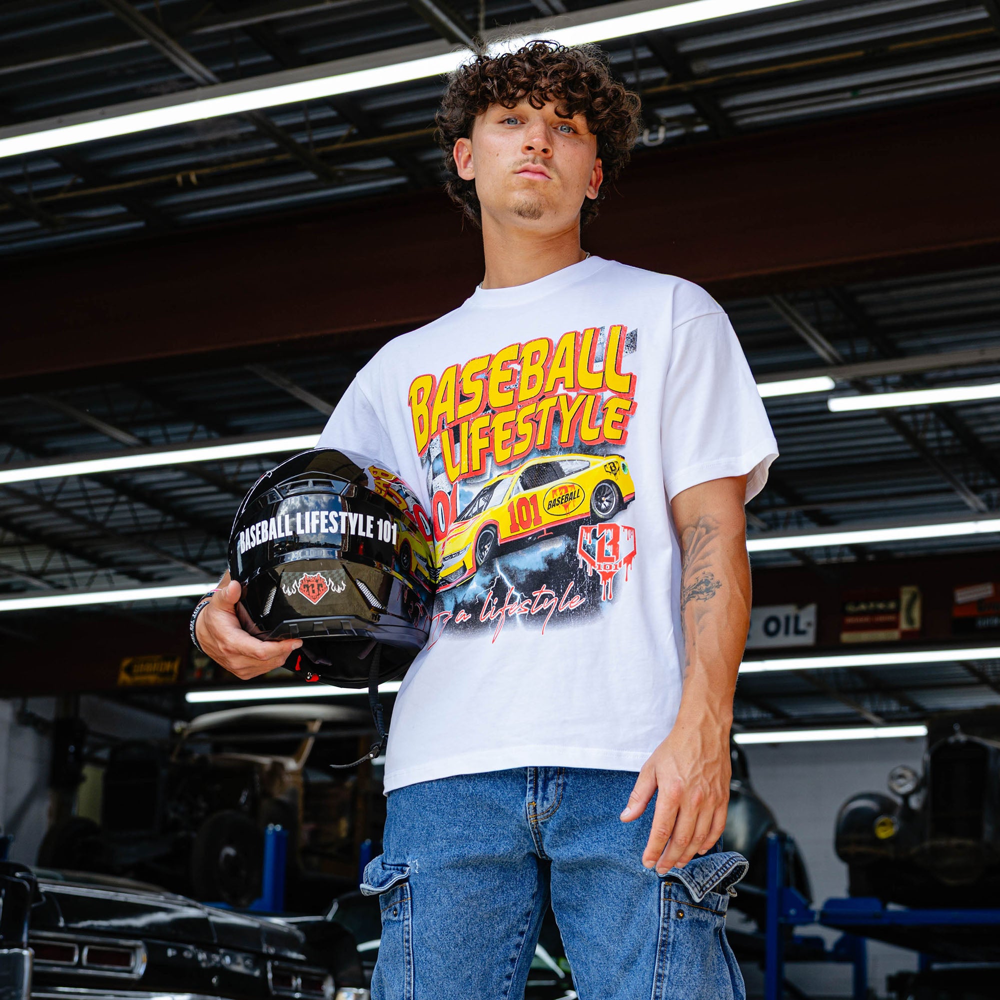 Young man holding racing helmet in vintage garage, wearing white Diamond Drift tee with yellow and red BL101 race car graphic and jeans.