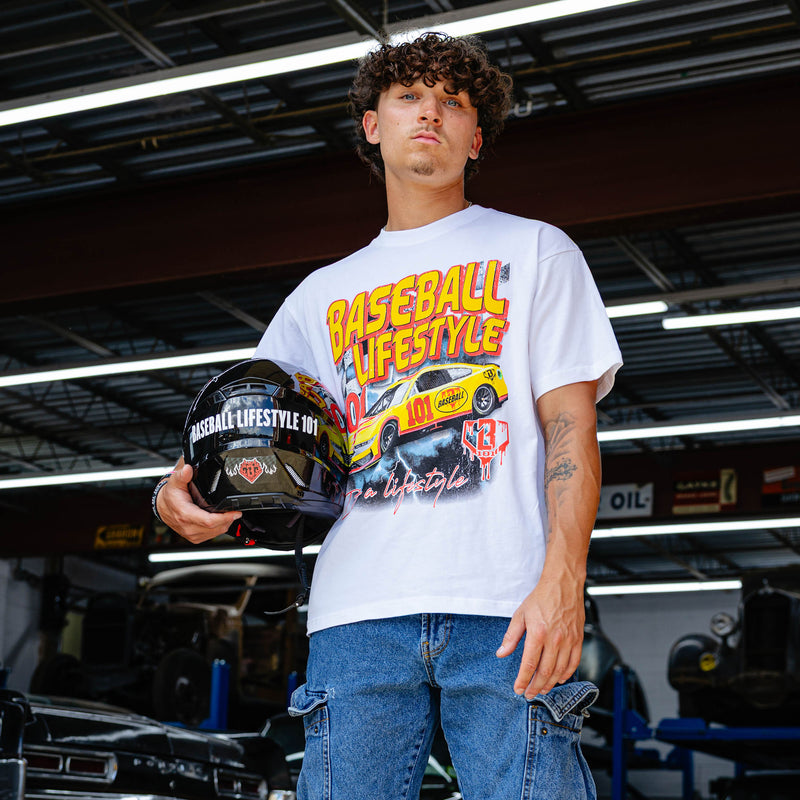 Young man holding racing helmet in vintage garage, wearing white Diamond Drift tee with yellow and red BL101 race car graphic and jeans.