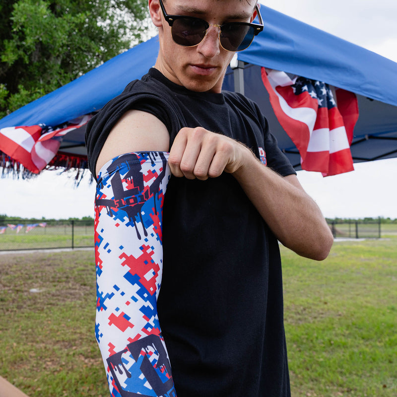 Adult male model flexing arm showing red white and blue digital camo arm sleeve with drip BL101 logo at outdoor baseball event