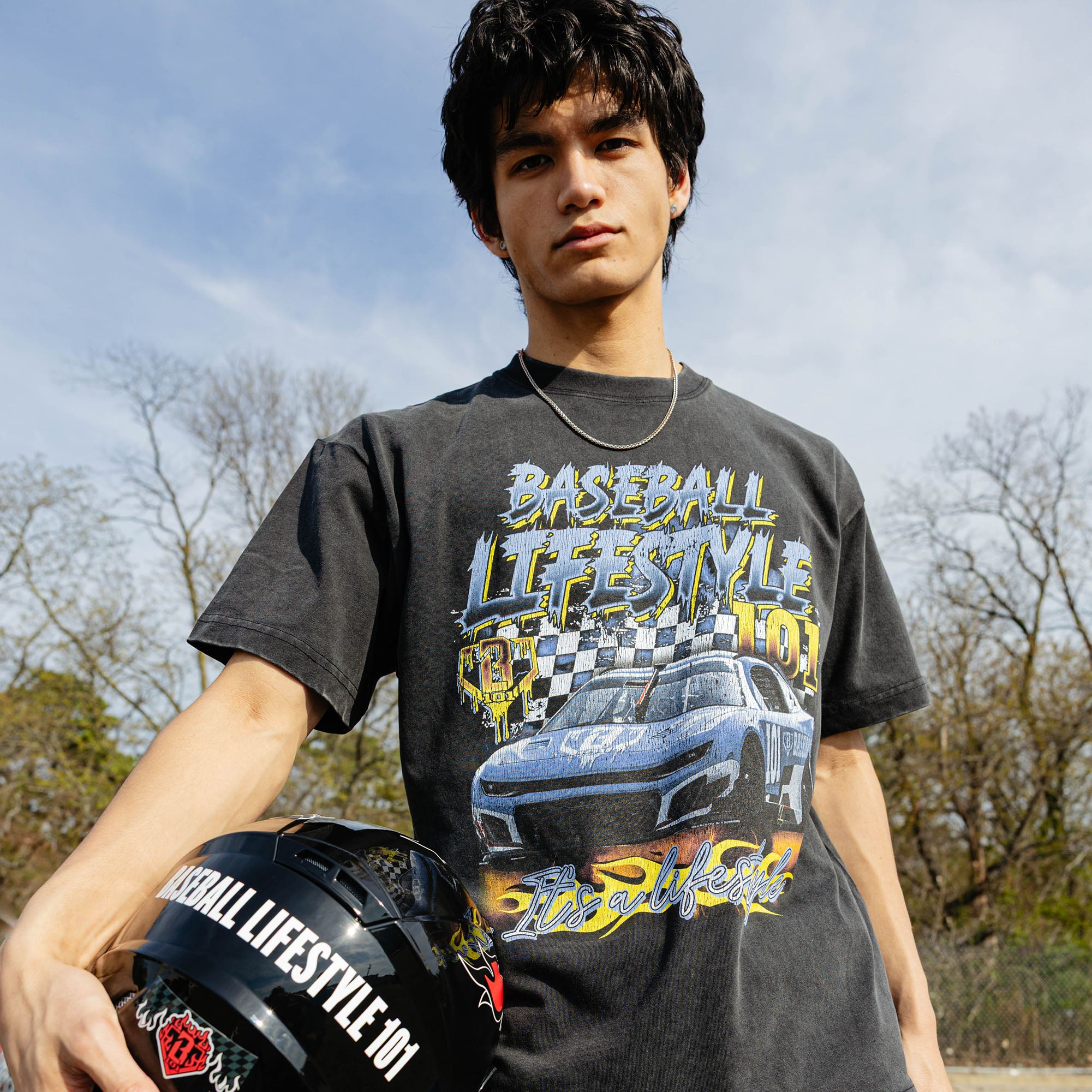 Young man facing forward holding Baseball Lifestyle 101 racing helmet, wearing washed black Fast Track Fielder tee with race car graphic, outdoors.