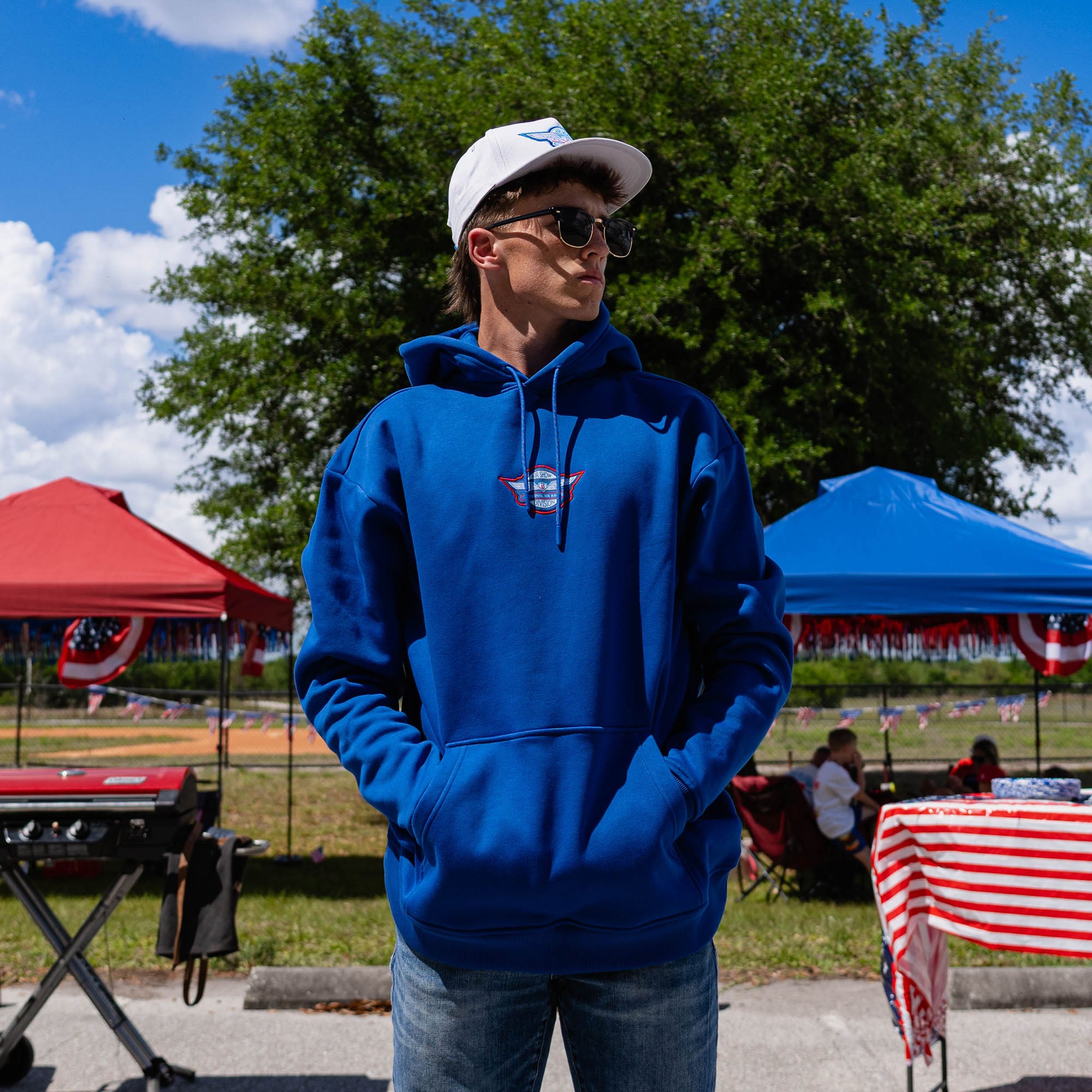 Front-facing adult male model wearing royal blue Fastball Flight hoodie with white snapback and sunglasses hands in pocket at outdoor baseball event