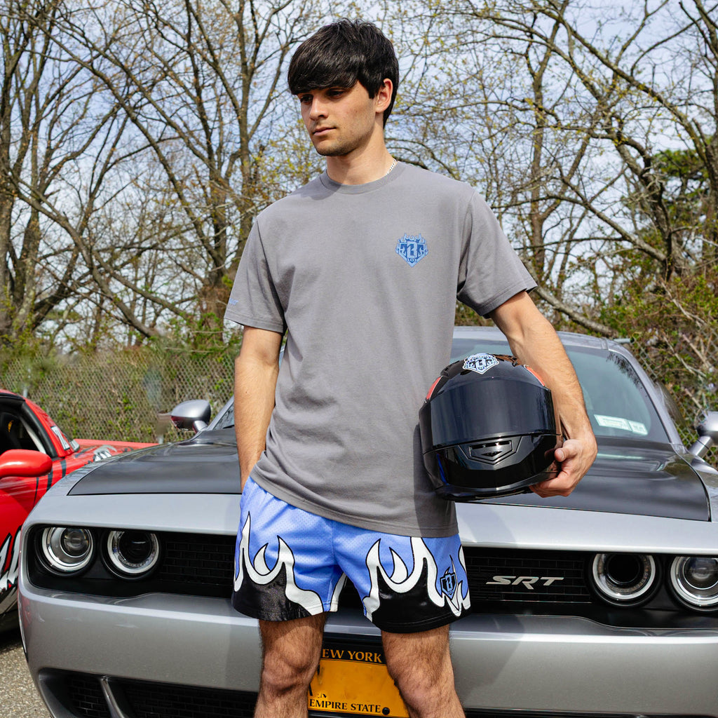 Young man holding racing helmet beside silver Dodge SRT Challenger, wearing gray Flame Thrower tee and blue flame mesh shorts.