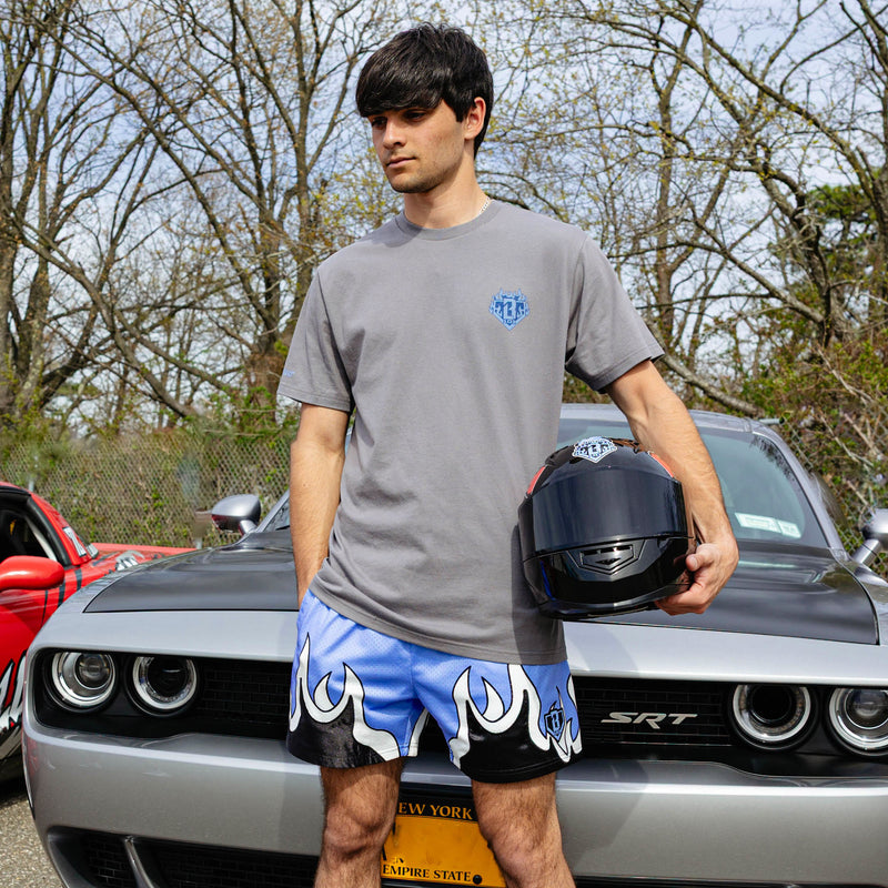 Young man holding racing helmet beside silver Dodge SRT Challenger, wearing gray Flame Thrower tee and blue flame mesh shorts.