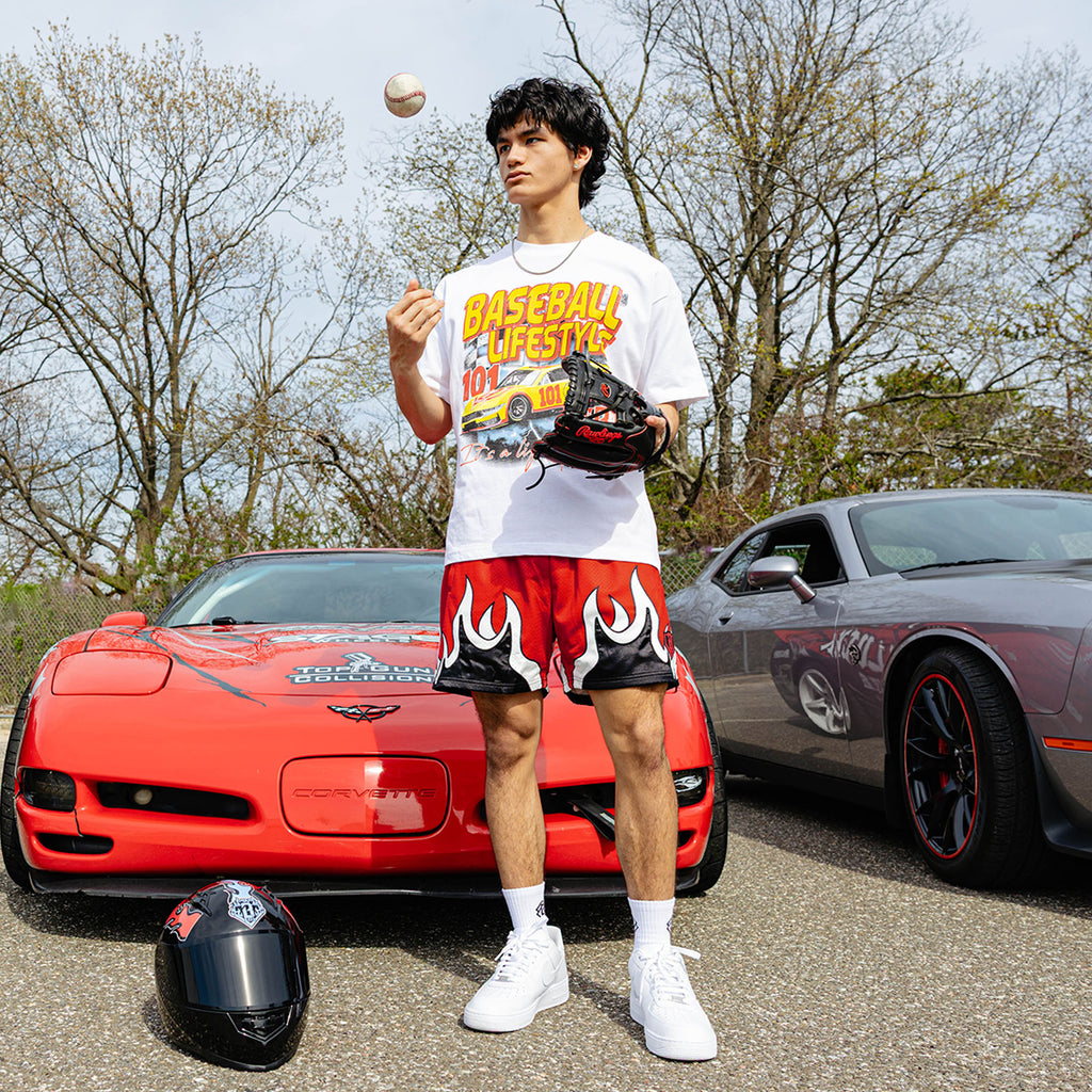 Young man tossing a baseball beside red Corvette and gray Dodge Challenger, wearing white BL101 race car tee and red flame mesh shorts, baseball glove in hand.