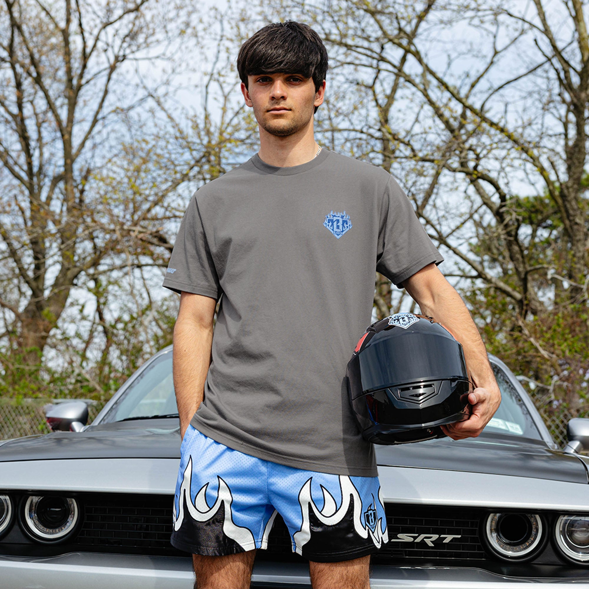 Young man standing in front of silver Dodge SRT Challenger holding racing helmet, wearing gray Flame Thrower tee with blue BL101 chest graphic and blue flame shorts.